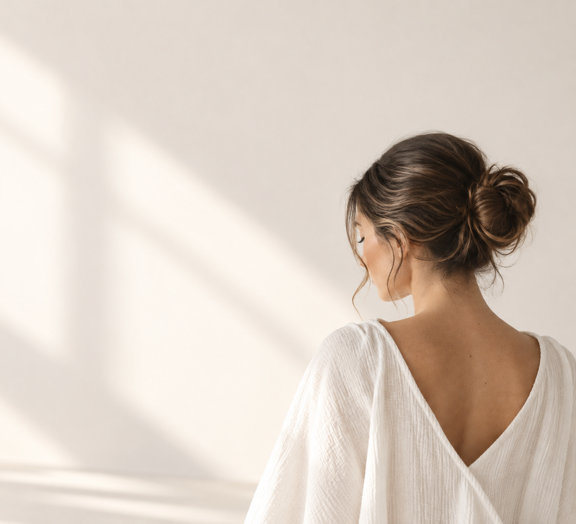 Woman with her hair in a loose bun, looking down in soft sunlight against a warm, neutral background.