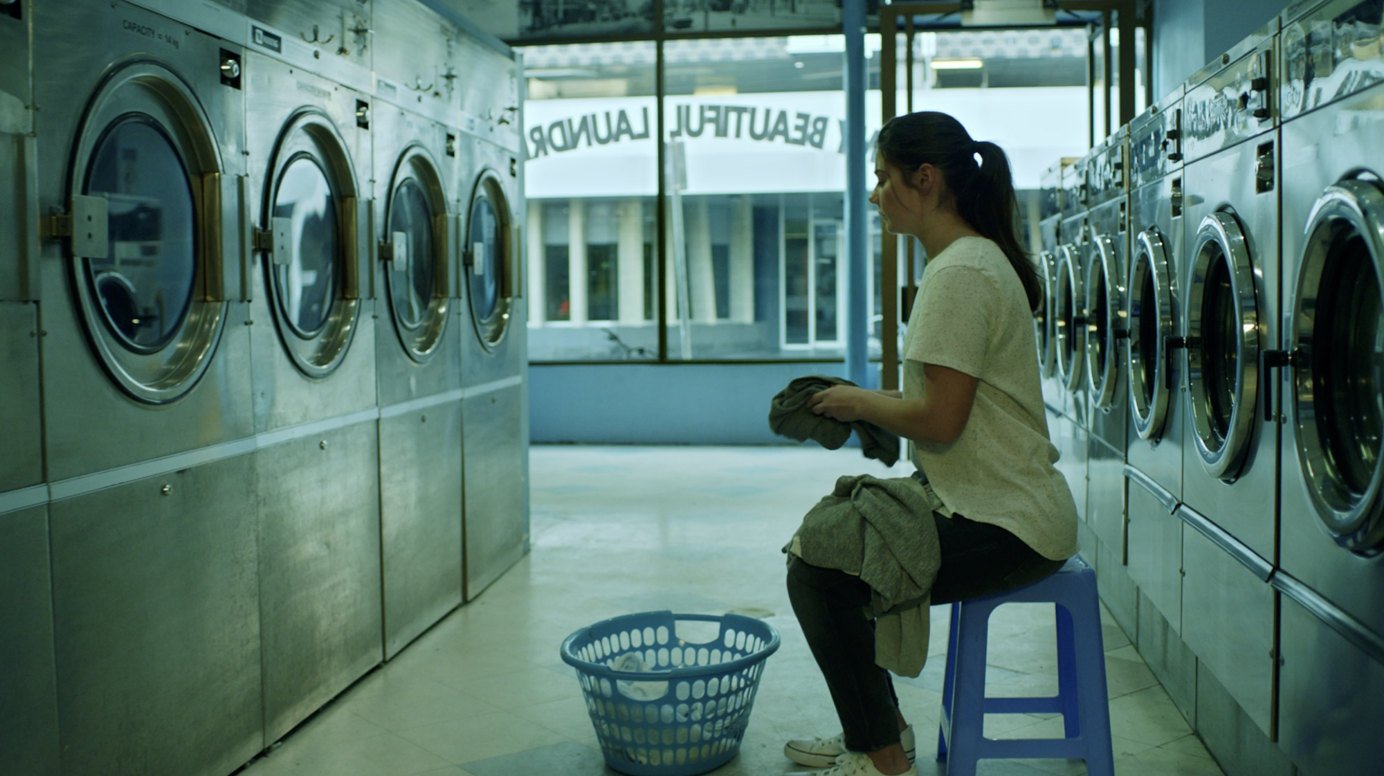 A young woman sits on a stool in a laundromat, holding clothes, surrounded by front-loading washing machines on both sides, with a laundry basket on the floor in front of her and a large window in the background.