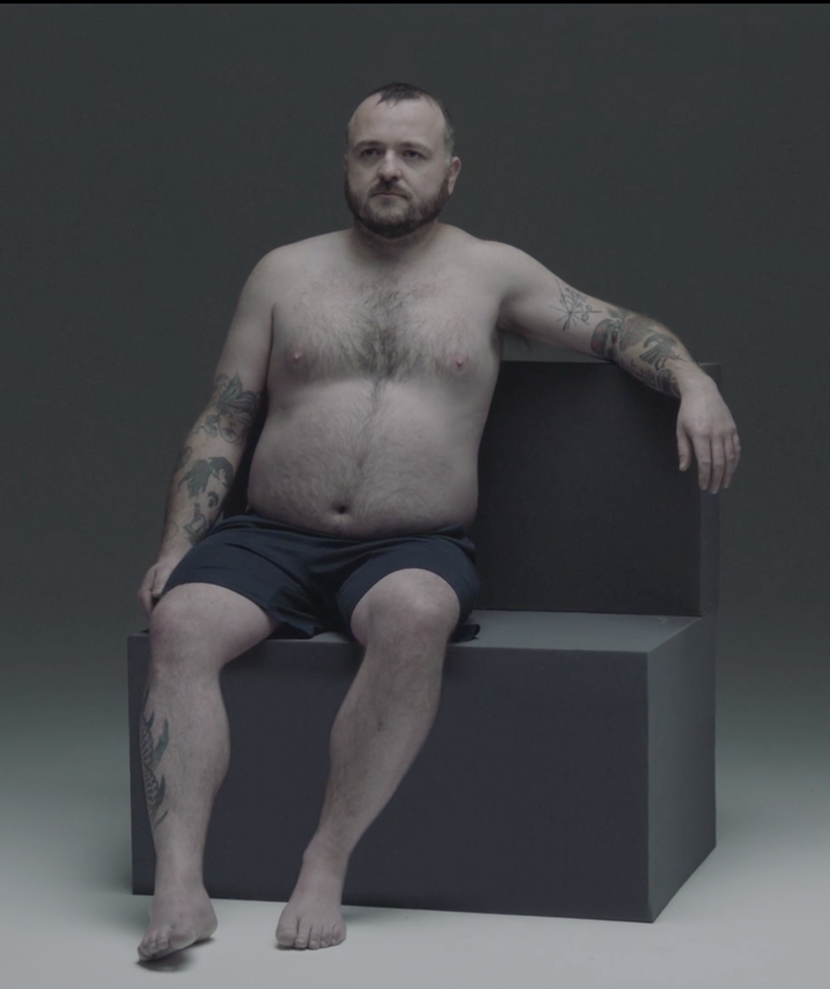 A shirtless man with tattoos on his arms and chest, sitting on a black bench against a dark gray background.