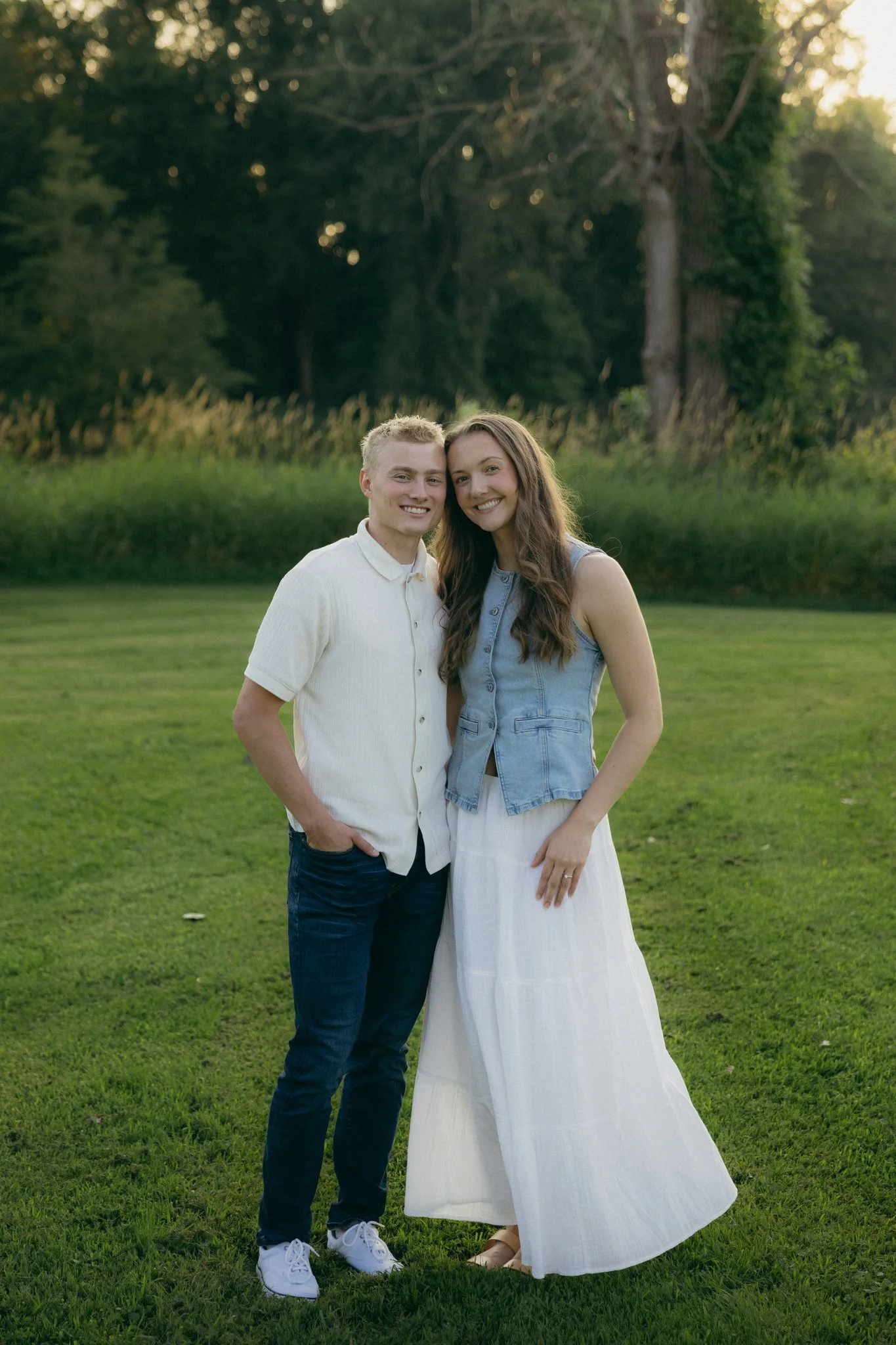 A young man and woman standing close together on a grassy field, smiling at the camera in an outdoor setting with trees and sunlight in the background.