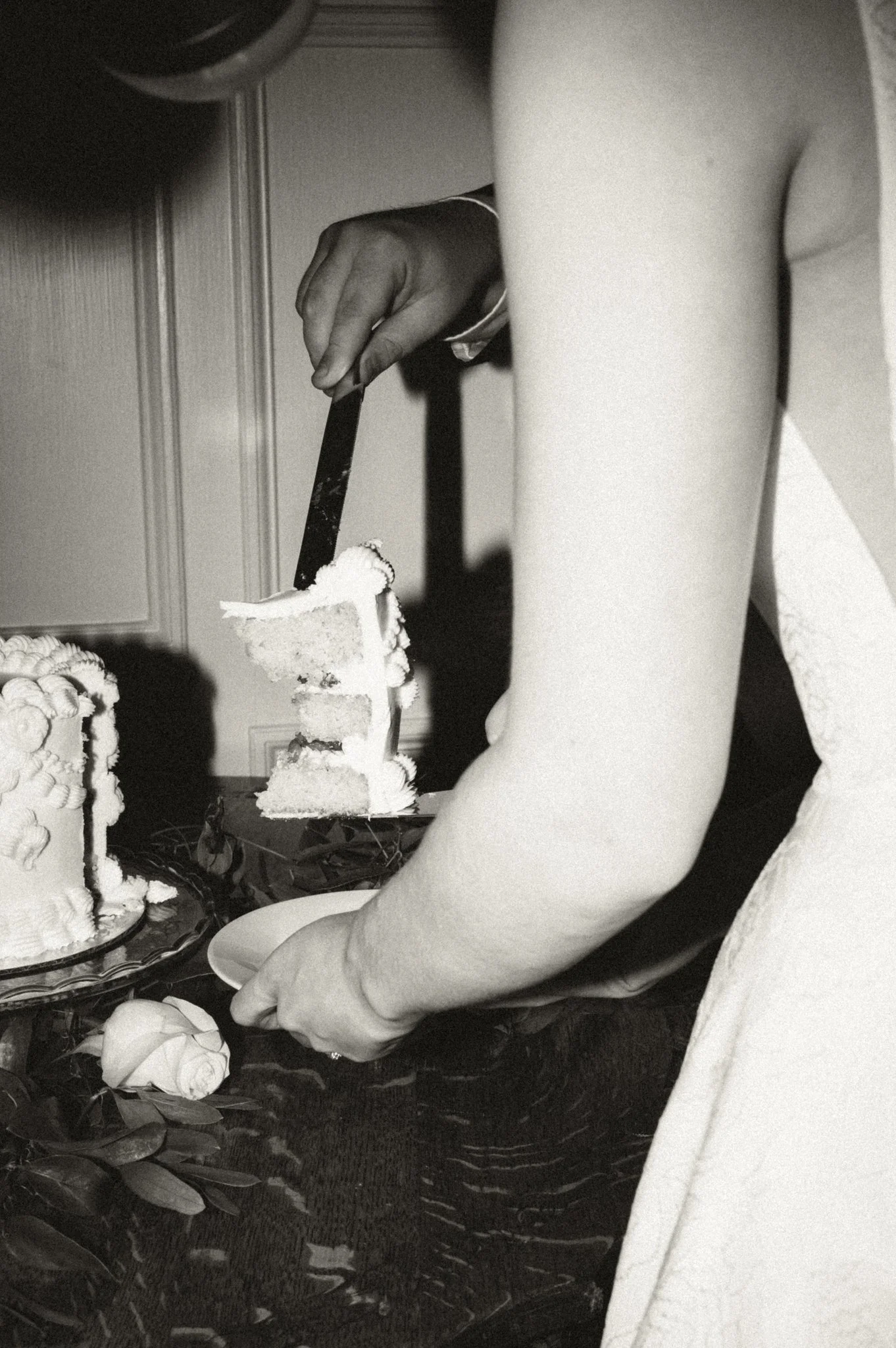 A person in formal attire is cutting a slice of wedding cake at a celebration event, with cakes and a flower on the table visible.