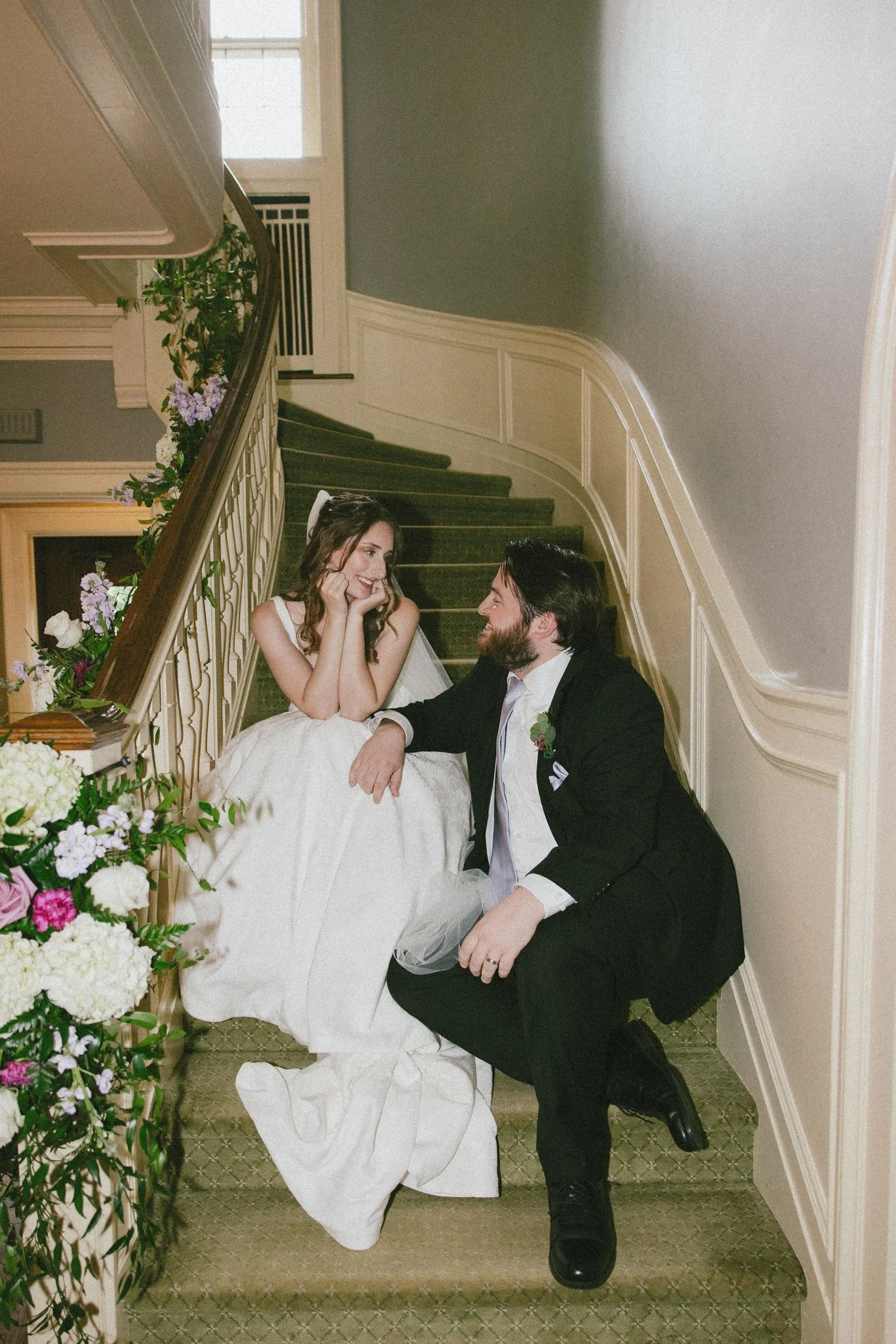 A bride and groom sitting on a staircase during their wedding, smiling and looking at each other, with flowers and greenery nearby.