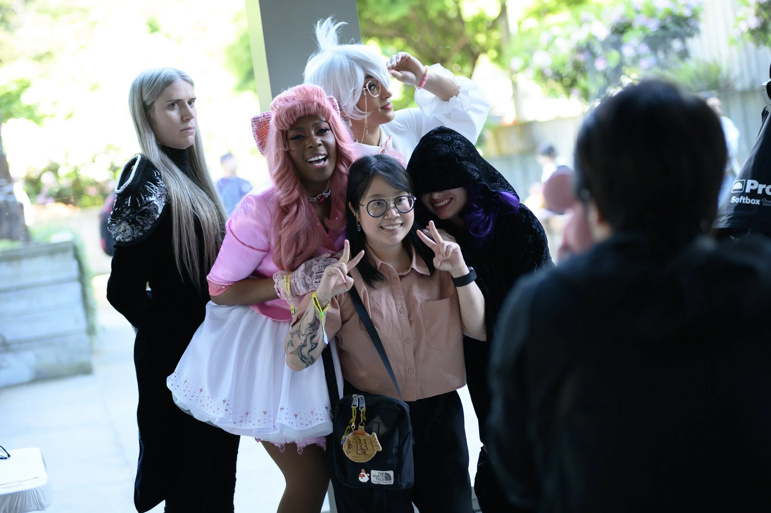 Group of five women cosplayers indoors, posing for a photo. They are dressed in colorful and elaborate costumes, with one making a peace sign, smiling at the camera.