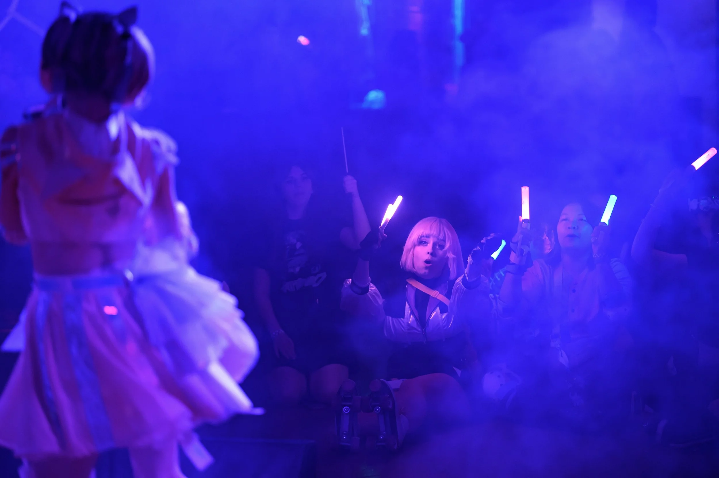 A group of young women with glow sticks in a dark, foggy environment, possibly at a concert or party, with blue and purple lighting, one woman sitting on the ground in the center looking surprised.
