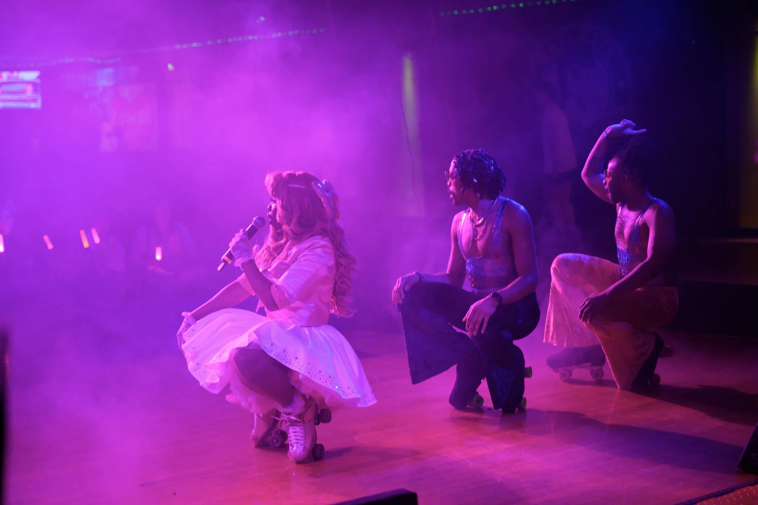 A woman with long blonde hair wearing a pink dress, holding a microphone, sitting on roller skates on a stage bathed in purple and pink lights. Two women with dark hair, also wearing roller skates and colorful outfits, sit on the edge of the stage wa