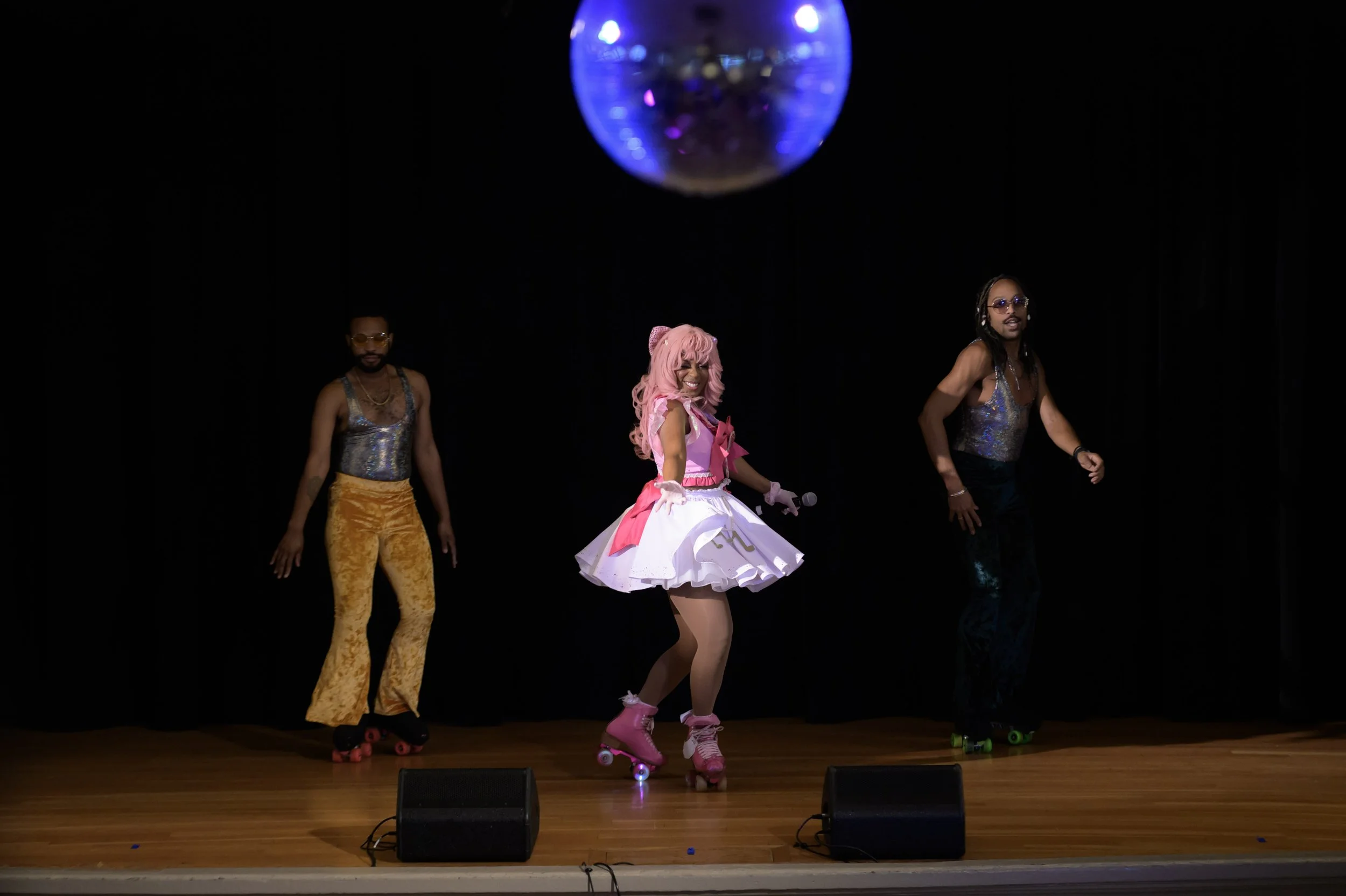 Three performers roller skating on stage with a disco ball overhead, black background, wooden stage floor, and two speakers in front.