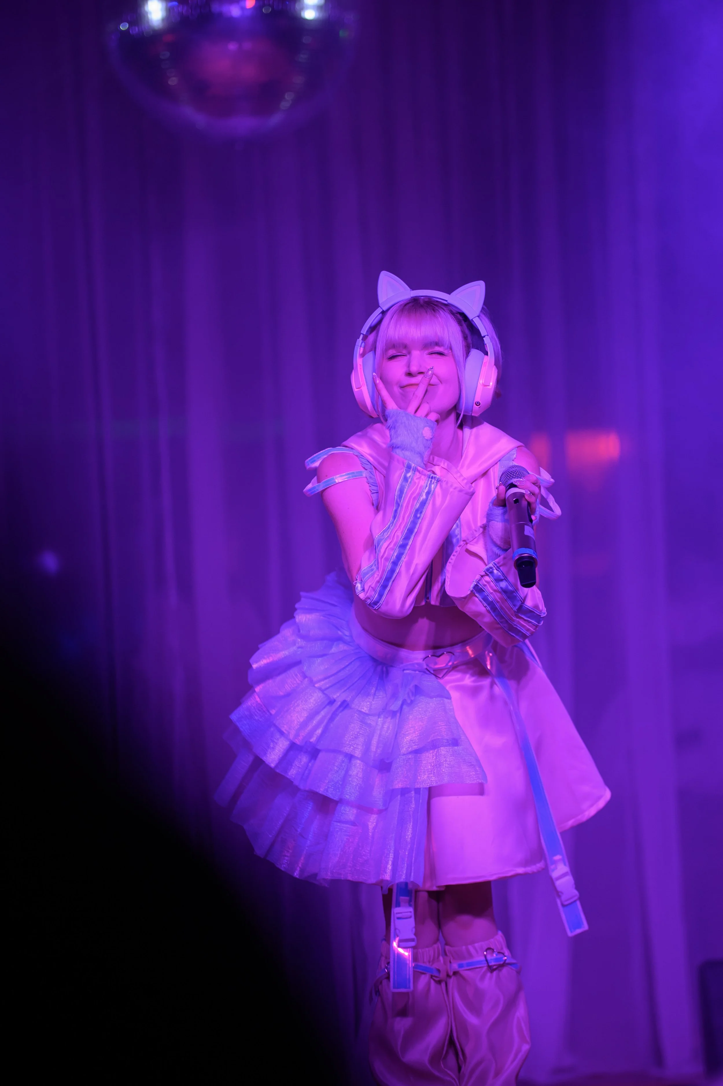 A young woman dressed in a pastel-colored, futuristic outfit with cat ear headphones, performing on stage with a microphone under purple lighting.