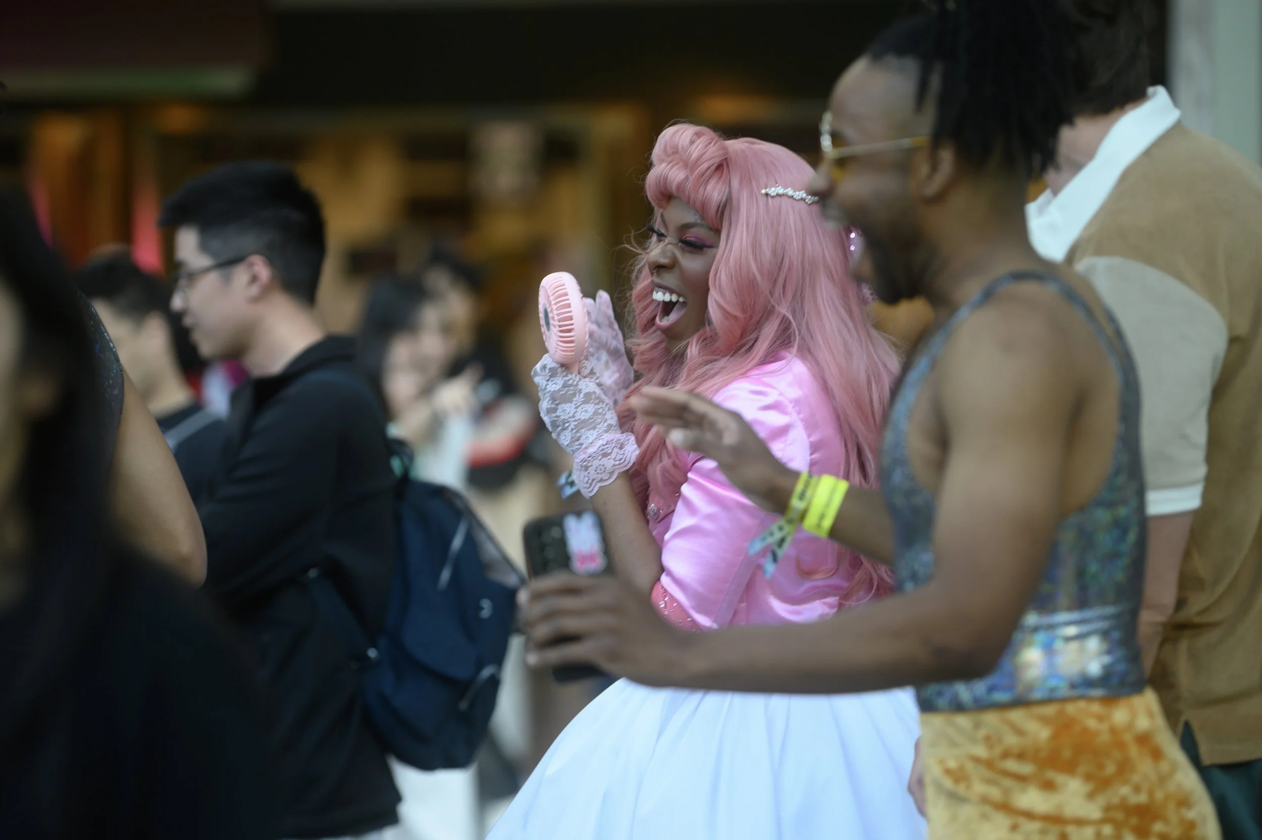 A woman with pink hair, dressed in a pink satin jacket and white skirt, laughs and plays with a small handheld fan at a crowded event, accompanied by others.