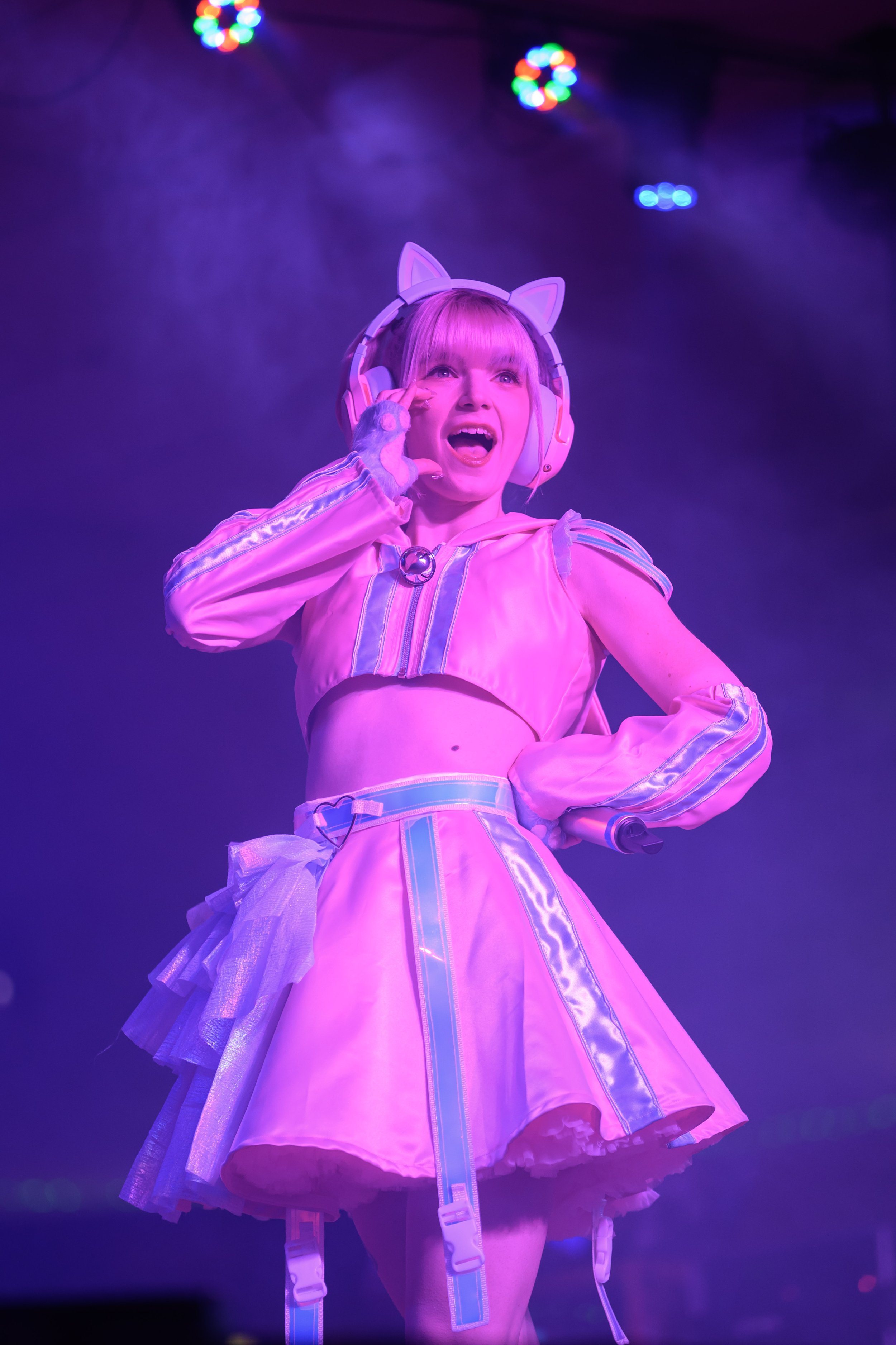 A young woman with blonde hair wearing a pink costume with iridescent accents and headphones with cat ears, performing on stage under colorful lights.