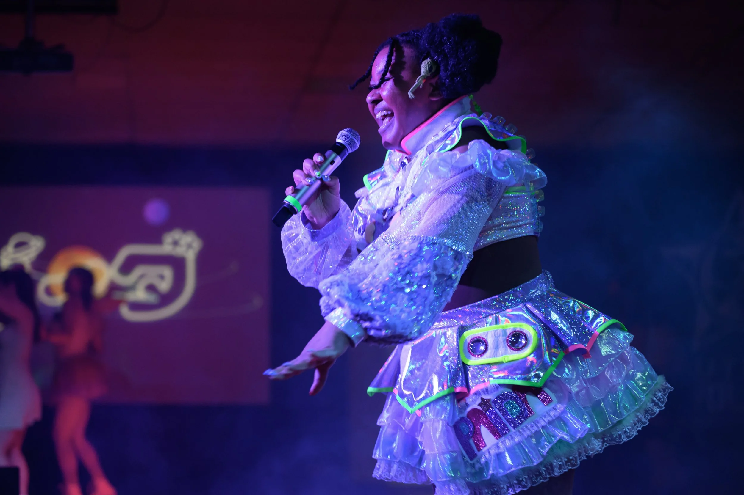 A performer singing into a microphone on a stage, dressed in a shiny, colorful costume with neon accents and the word 'PAiDA' on the skirt, under vibrant stage lighting.