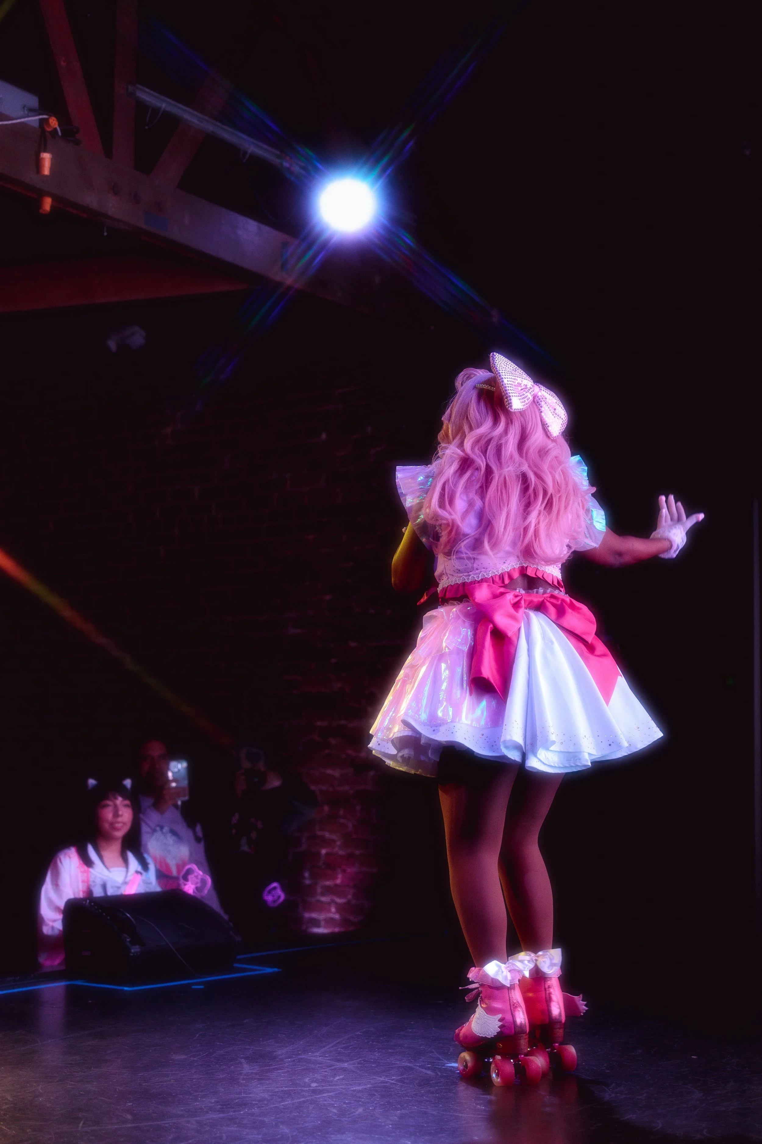 A girl with pink hair in a colorful, frilly costume with roller skates performing on stage; audience members watching.