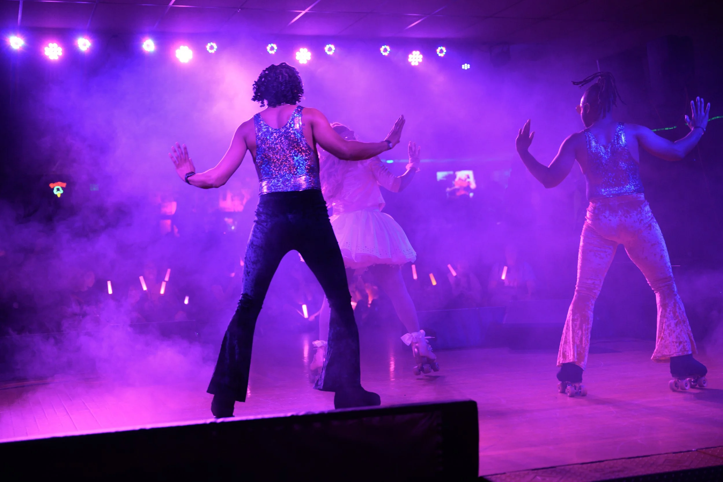 Three people roller skating on a stage with purple and pink lighting and fog, performing dance moves.