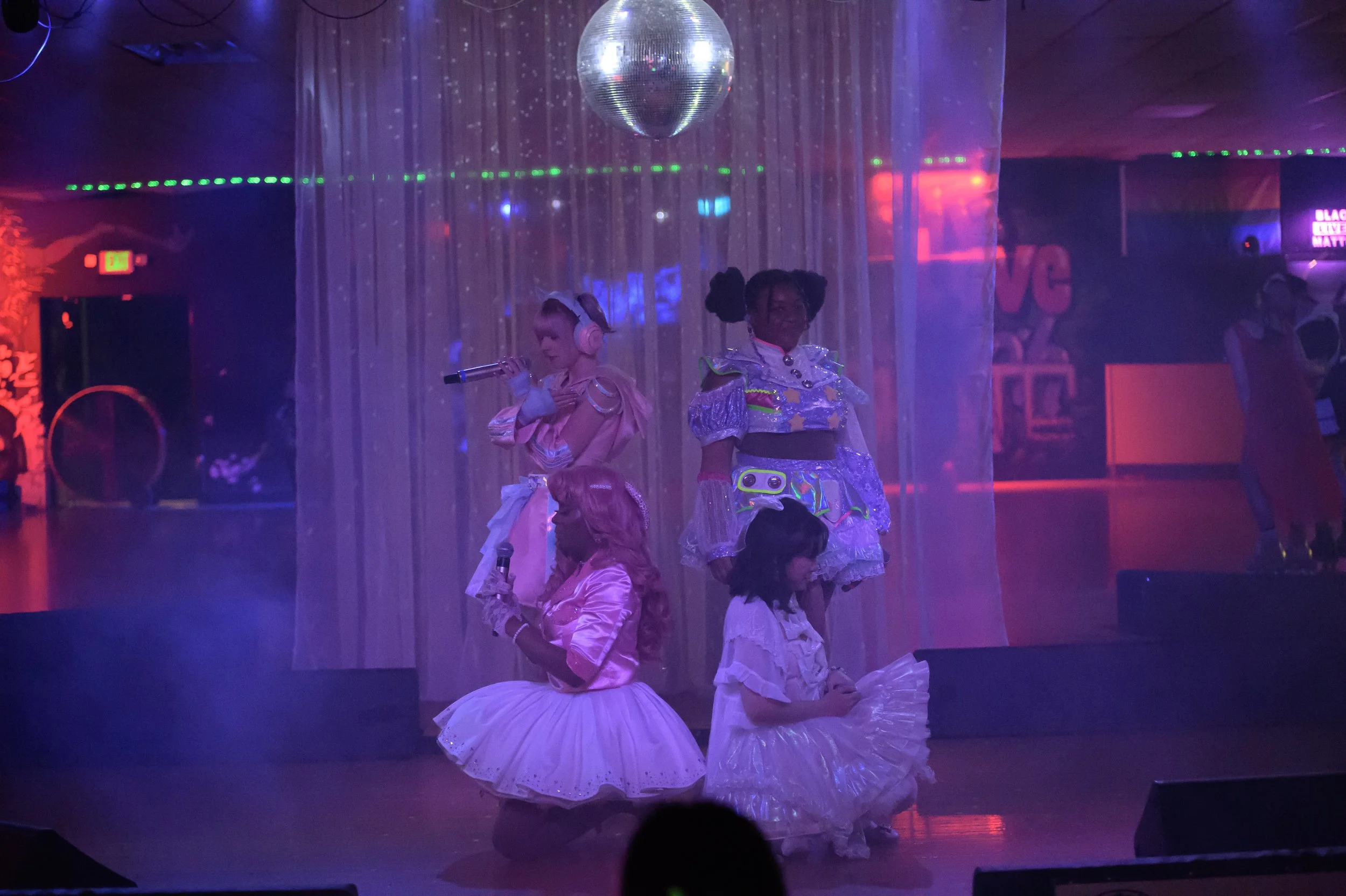 Four women dressed in colorful, futuristic costumes on a dimly lit stage with a disco ball overhead, performing in a roller rink setting.