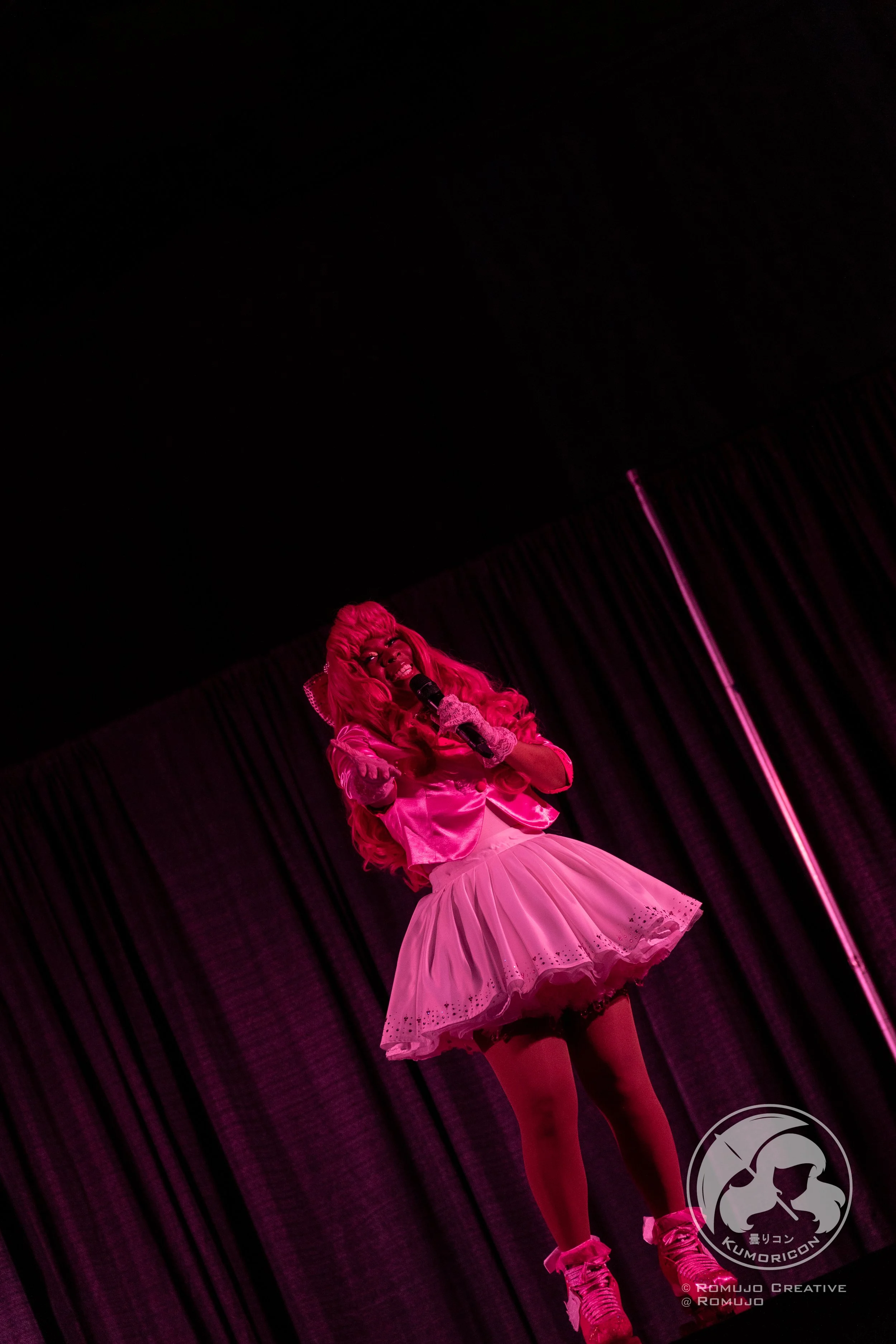 Performer dressed in pink, with long curly pink hair, singing into a microphone on stage with dark curtains in the background, illuminated by pink lighting.