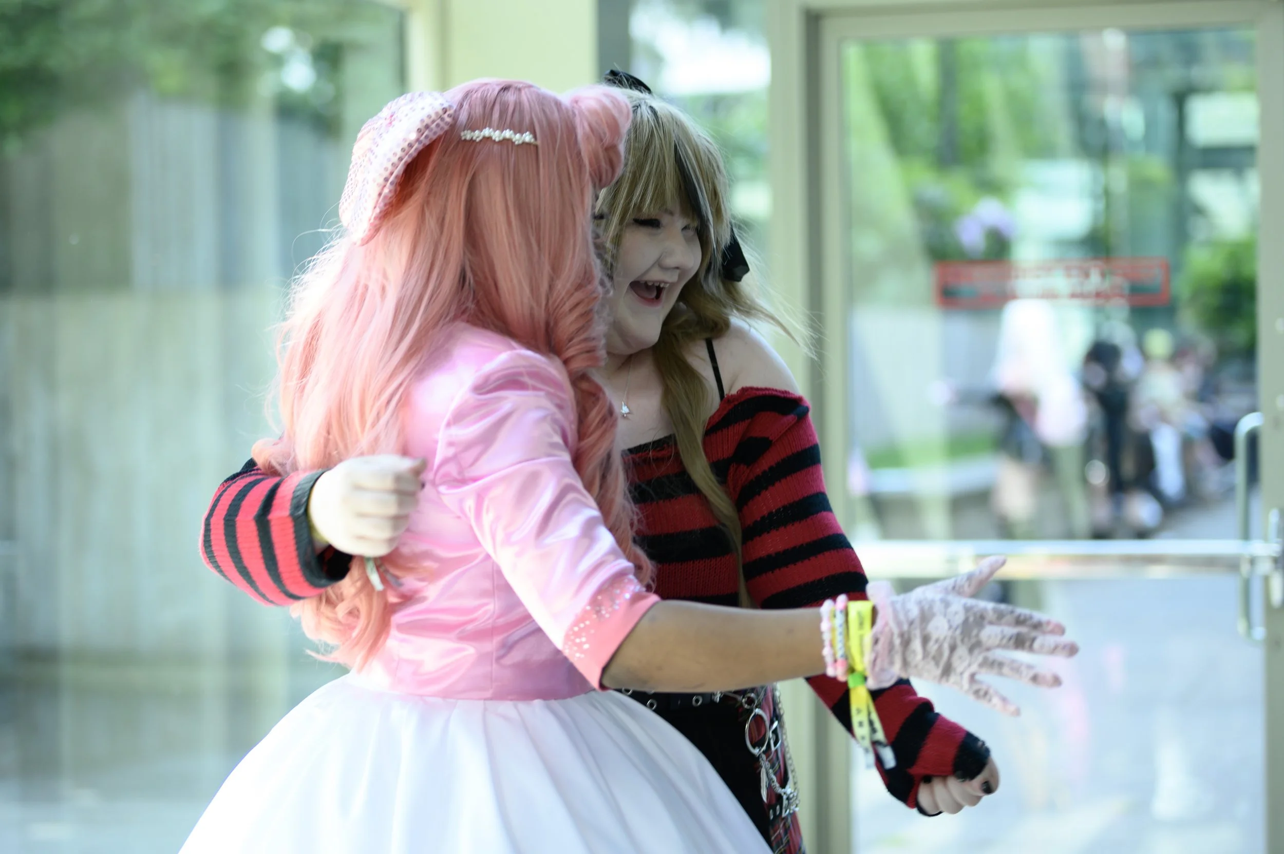 Two women, one in a pink dress and pink wig, and the other in a red and black striped sweater, share a joyful moment together, smiling and pointing at something outside a glass building.