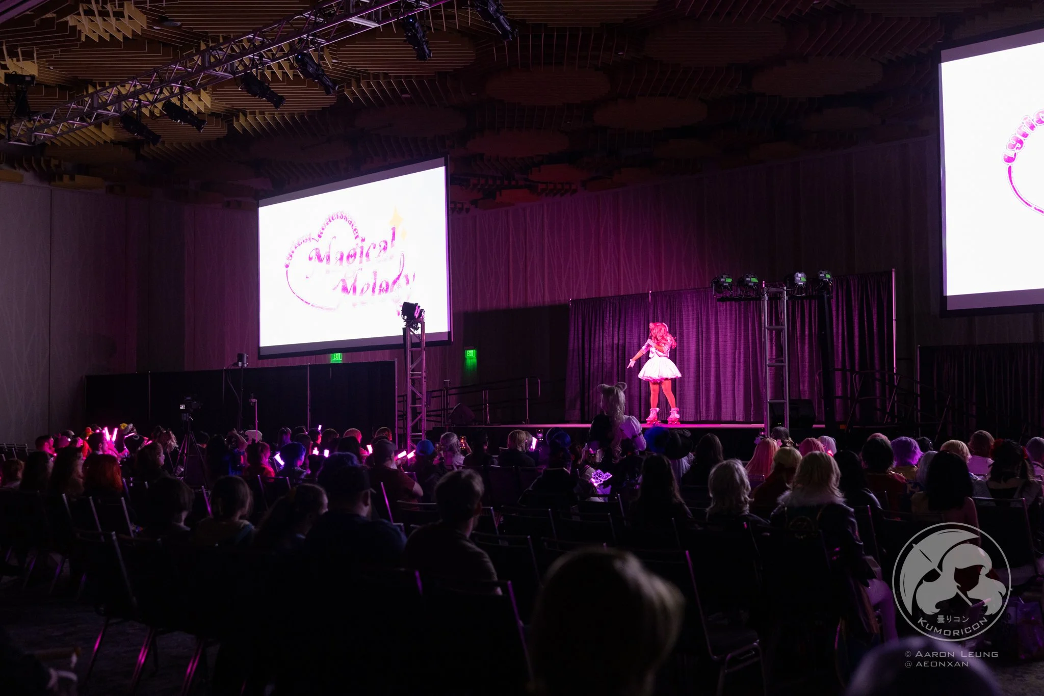 A performer dressed in pink on stage at the Kumoricon 2024 , with large screens displaying 'MAGICAL MELODY' and an audience watching under purple and pink lighting.