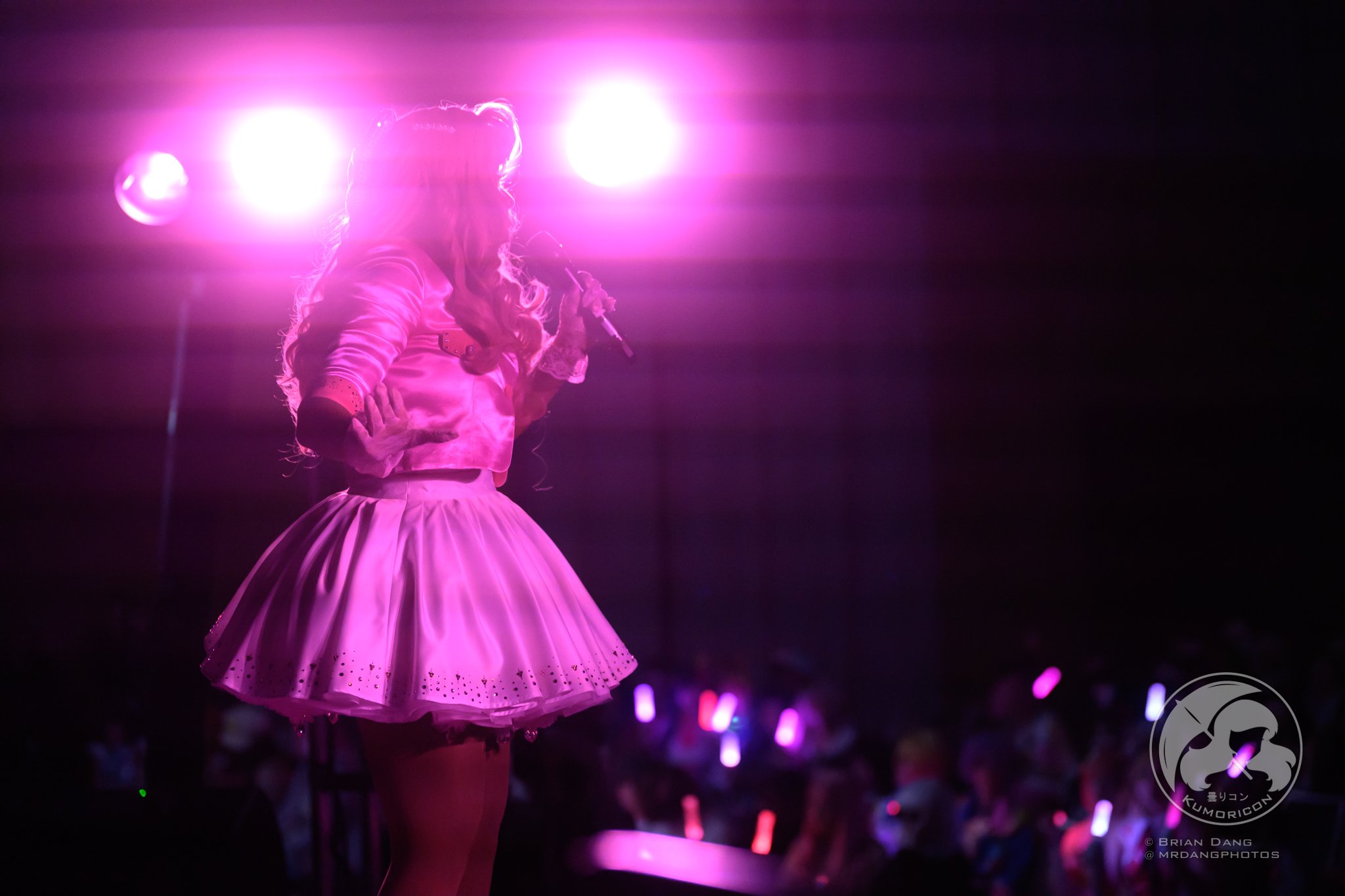 Female performer on stage holding a microphone, illuminated by bright pink and purple stage lights, with an audience holding glow sticks in the background.