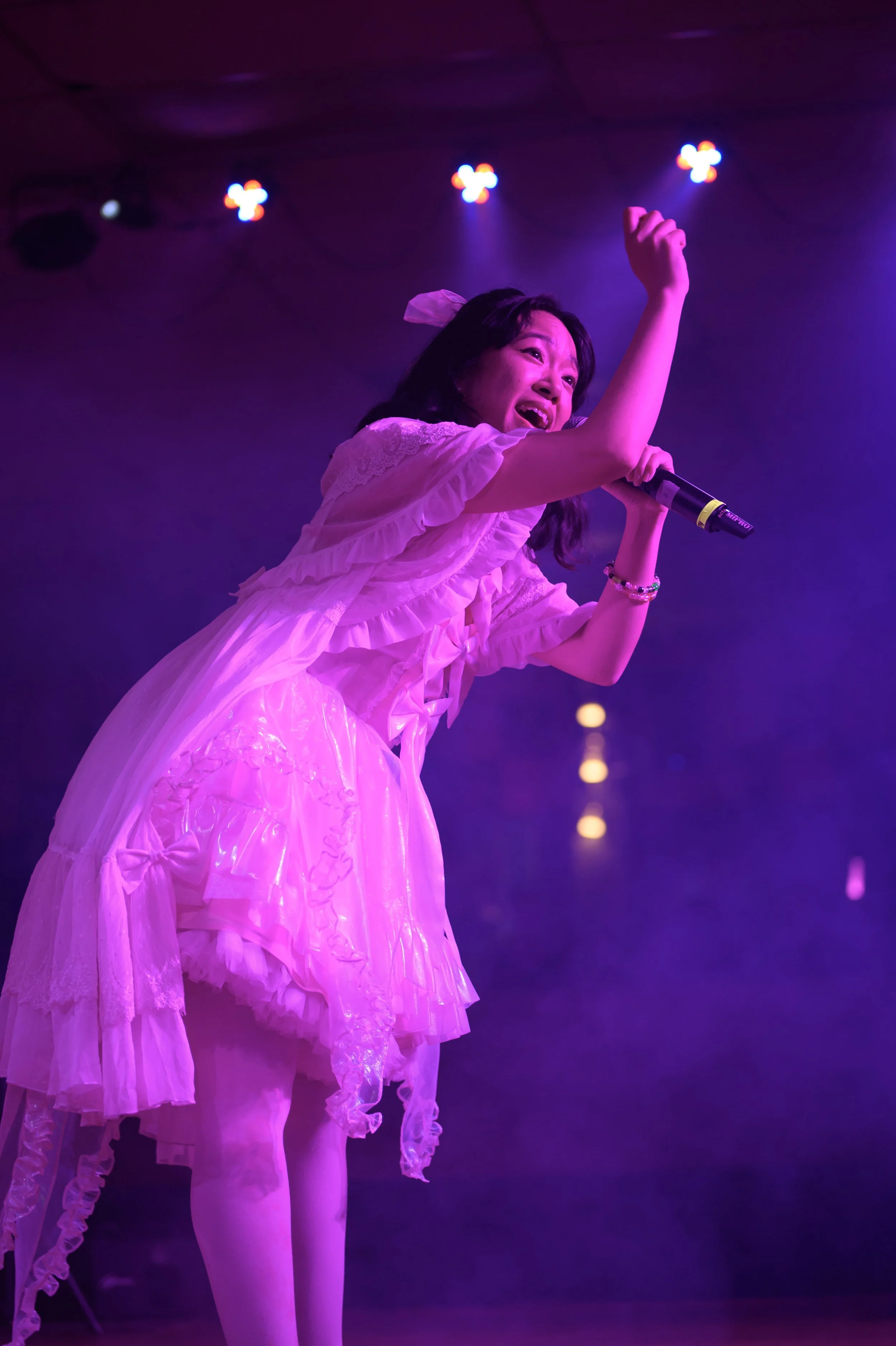 A woman wearing a pink dress singing into a microphone on stage under purple lights.