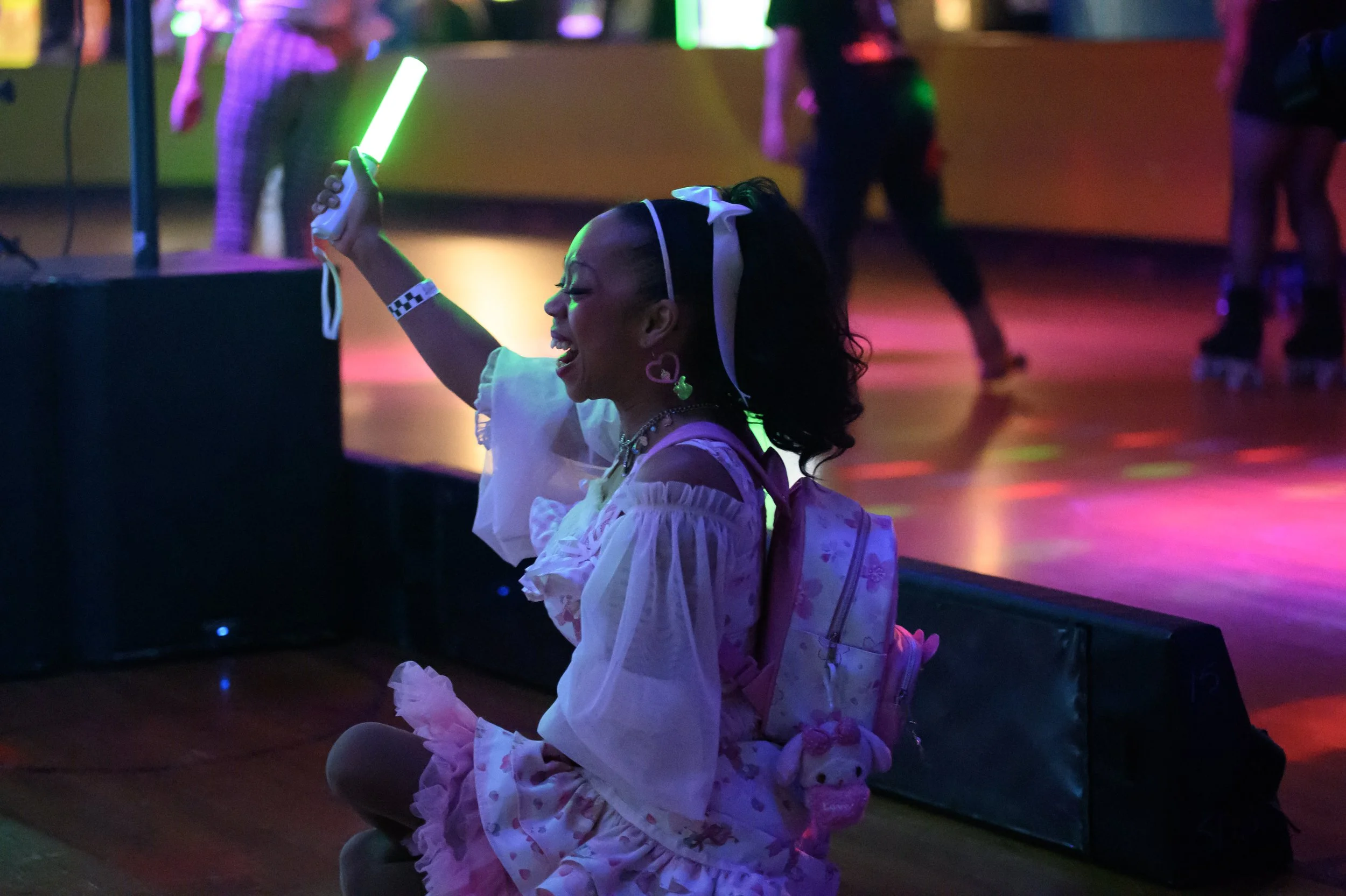 A woman at a roller skating rink, kneeling on the floor and smiling while holding a glowing green light stick. She is dressed in a pastel-colored, ruffled dress with a backpack, and has earrings, a headband, and a plush doll attached to her backpack.