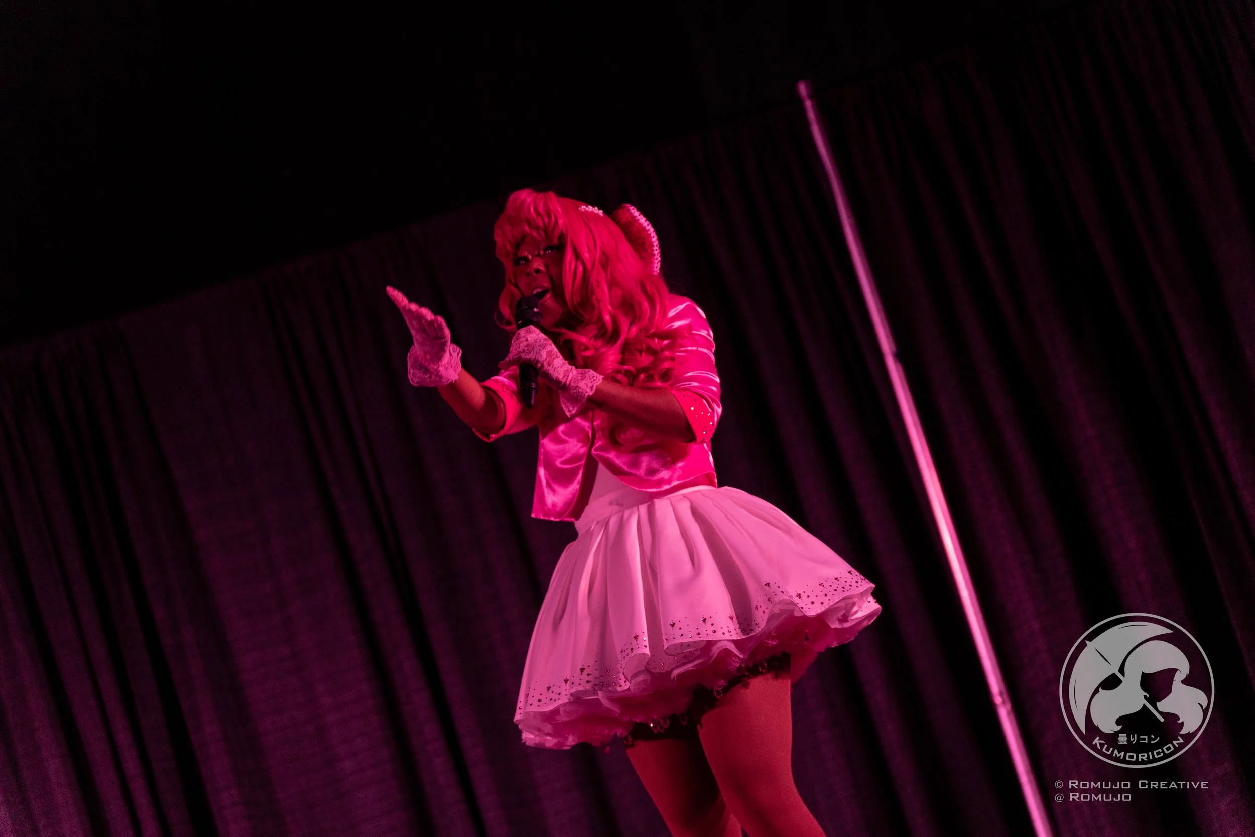 Person dressed in a pink costume performing on stage with a black curtain in the background and a stage pole to the right.