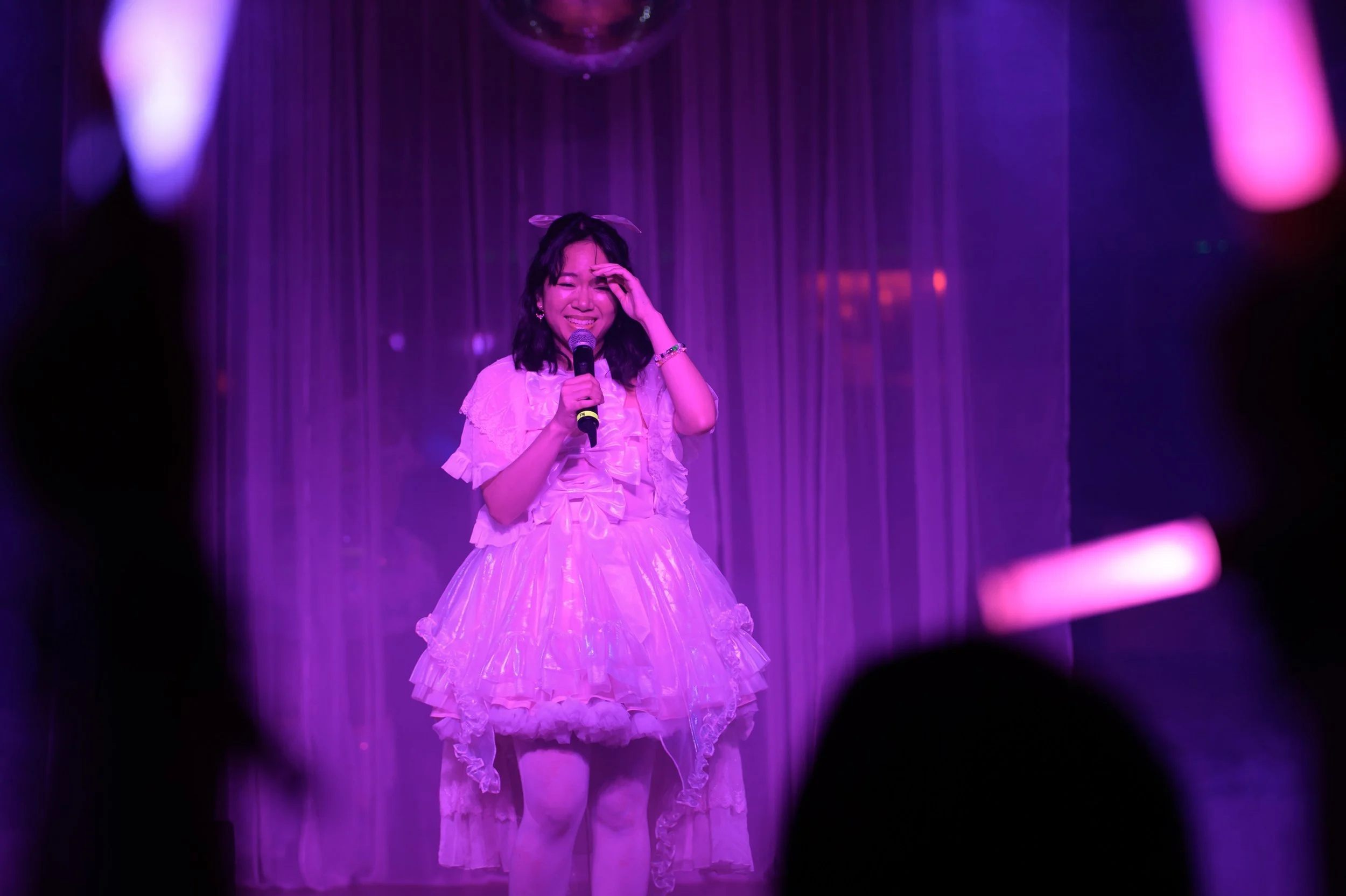 A young woman in a pink, frilly dress holding a microphone on stage, smiling and wiping her eye, with purple stage curtains in the background.