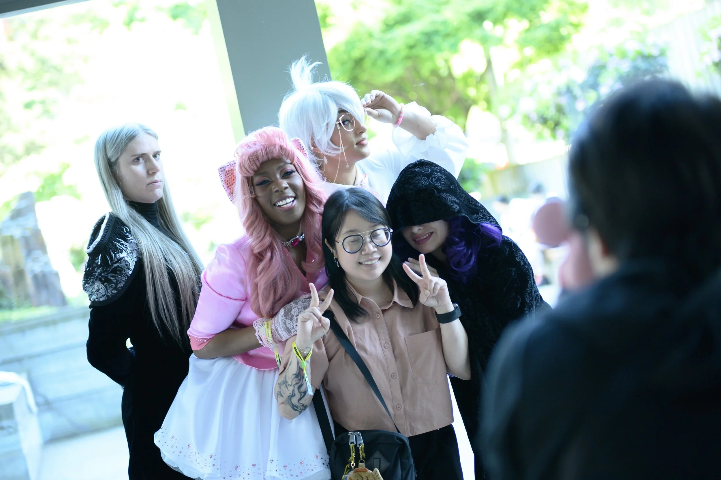 Group of five diverse women posing for a photo outdoors during the day, dressed in colorful and quirky outfits, with smiles and peace signs, with a person taking their picture.