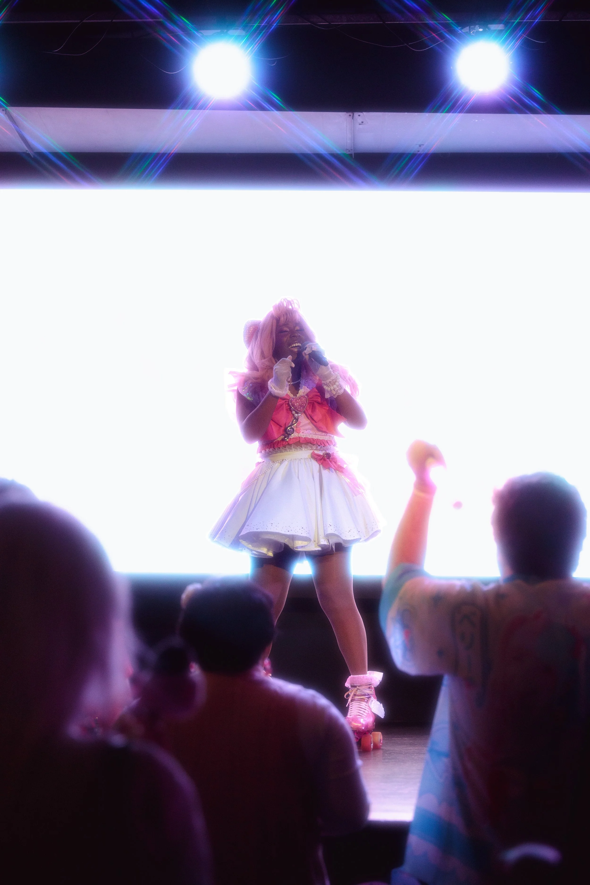 A performer dressed in a colorful, whimsical outfit with pink hair singing on stage while audience members watch.