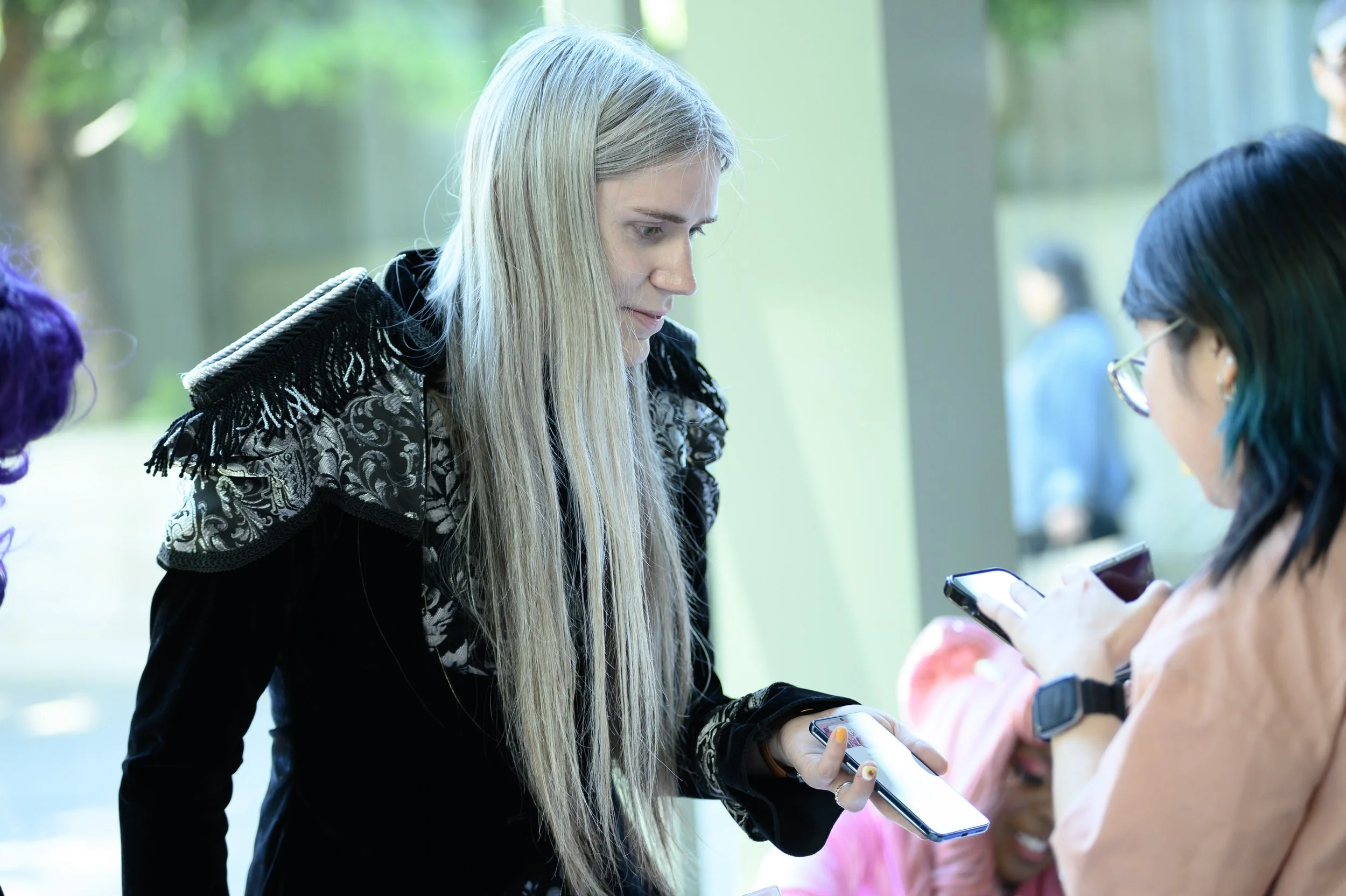 A woman with long blonde hair, wearing a black jacket with ornate silver embroidery, leaning forward and showing a smartphone to another woman with glasses and dyed teal hair, who is also using a smartphone.