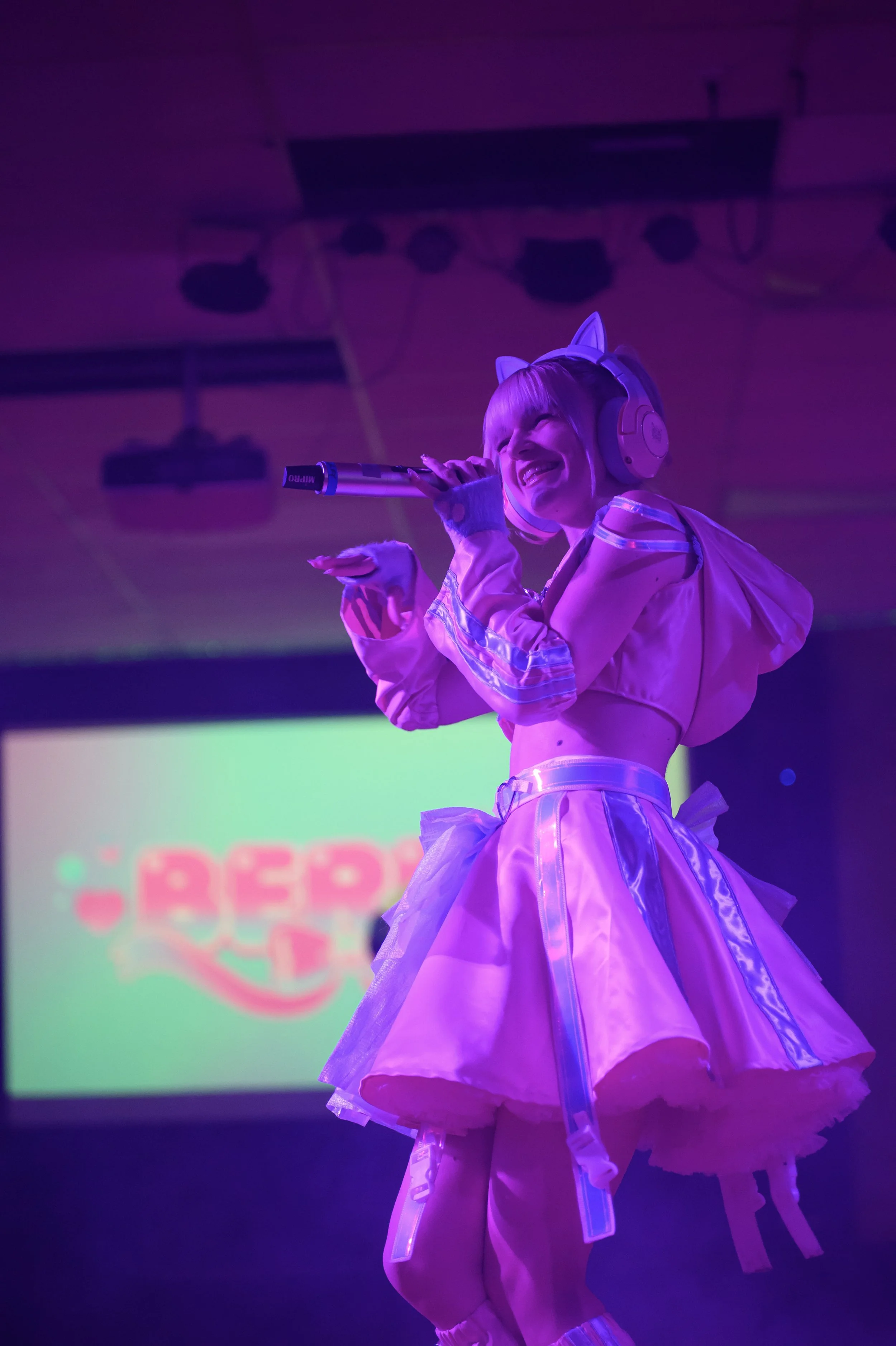 A person dressed in a pink, futuristic costume with cat ears, singing into a microphone on stage with purple lighting and a blurred screen in the background.