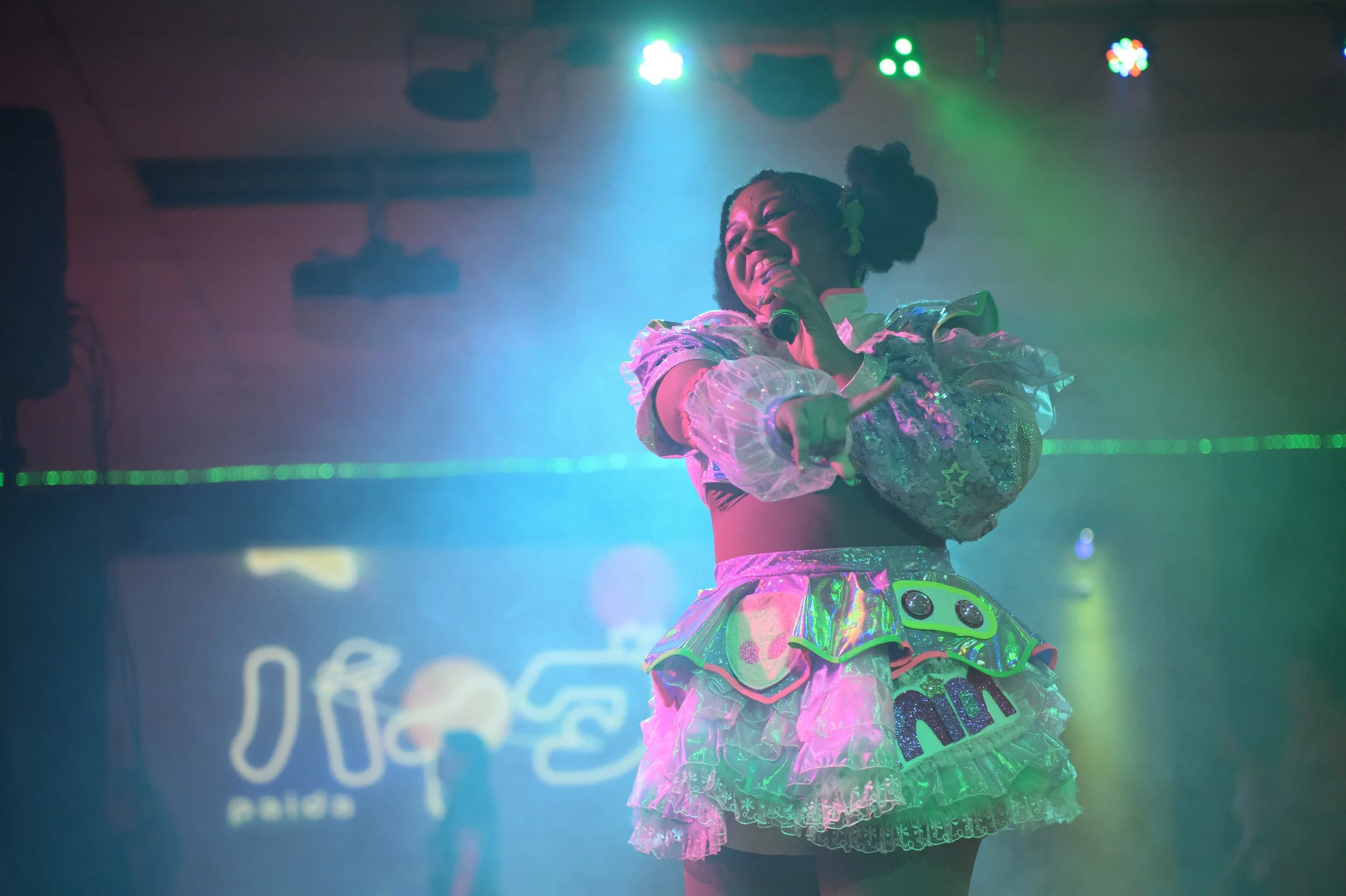 A performer on stage wearing a colorful, shiny outfit with puffy sleeves, a layered skirt with the word 'MISSION,' and pigtails, smiling and holding a microphone amid vibrant blue and green stage lights.