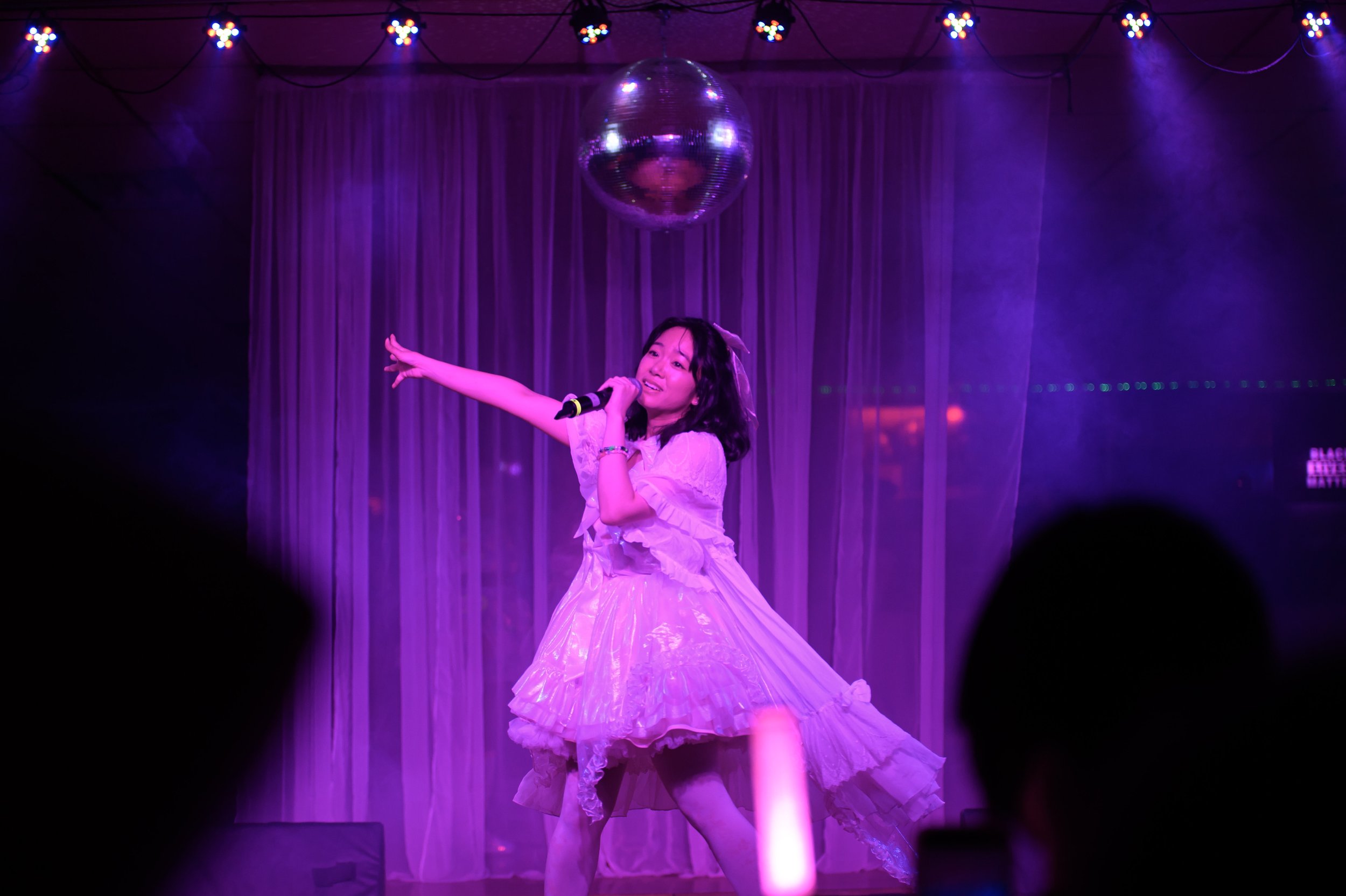 A female singer performs on a stage with purple lighting, wearing a pink princess-style dress with puffed sleeves and ruffles, holding a microphone, under a disco ball with purple curtains in the background, and audience silhouettes in the foreground
