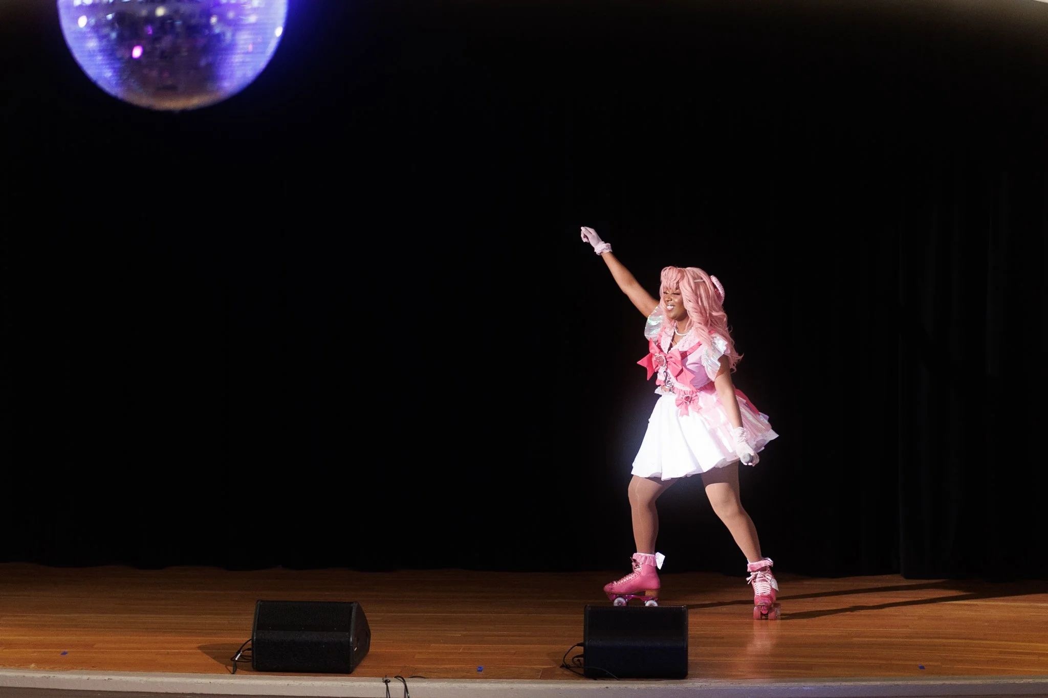 A woman dressed in pink and white roller skating costume, with pink hair, performing on stage with black curtains and a disco ball overhead.