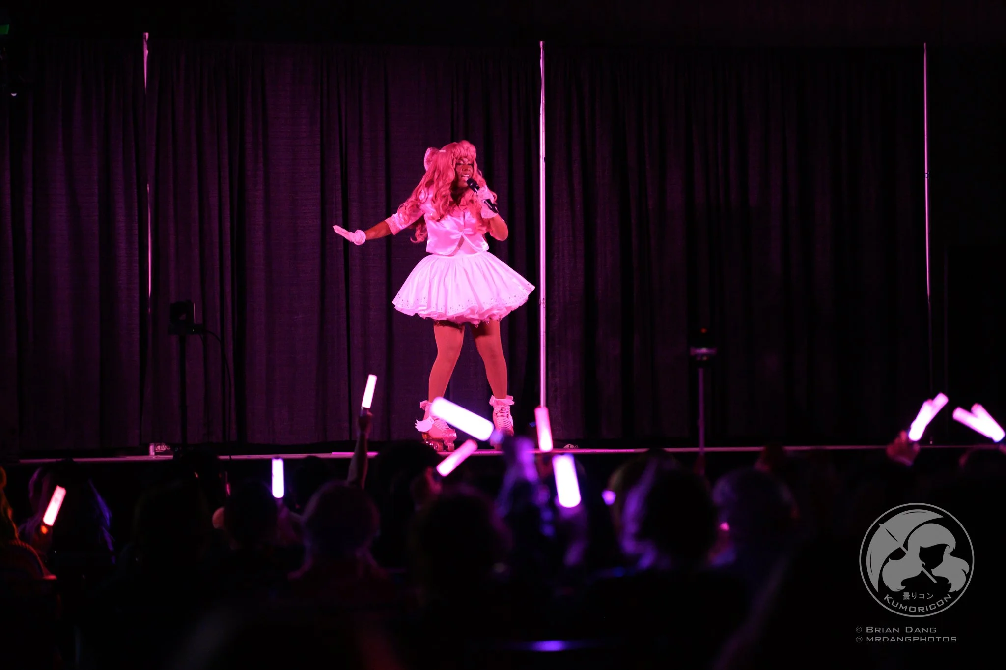 A performer dressed in a pink costume with a tutu skirt, long pink hair, and roller skates, singing into a microphone on a stage with a dark curtain backdrop, with an audience holding glow sticks in the foreground.