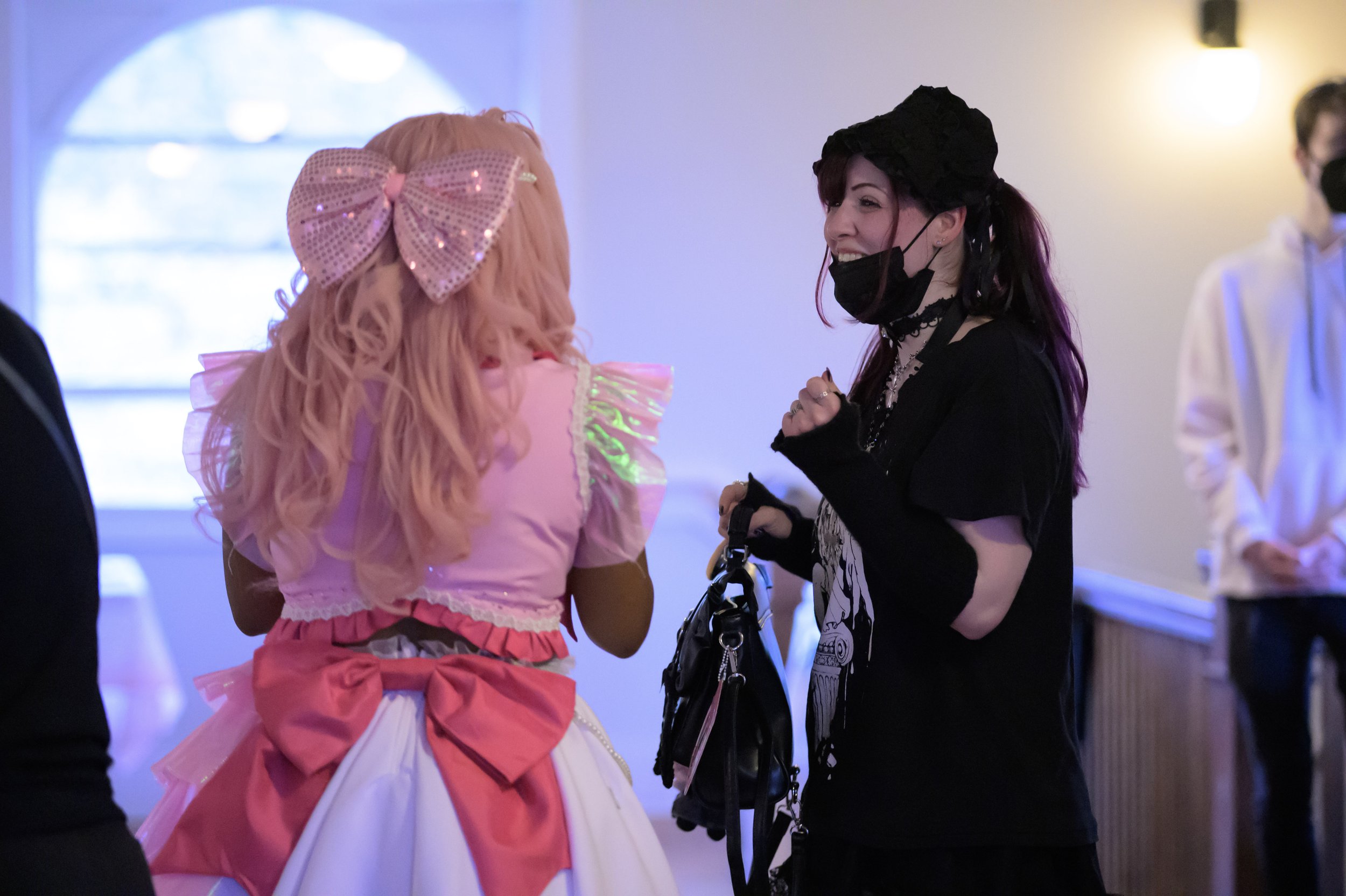 Two women wearing masks are talking indoors. One woman has pink hair with a large pink bow and a pink and white dress. The other woman has dark hair with purple streaks, dressed in black, holding a bag.