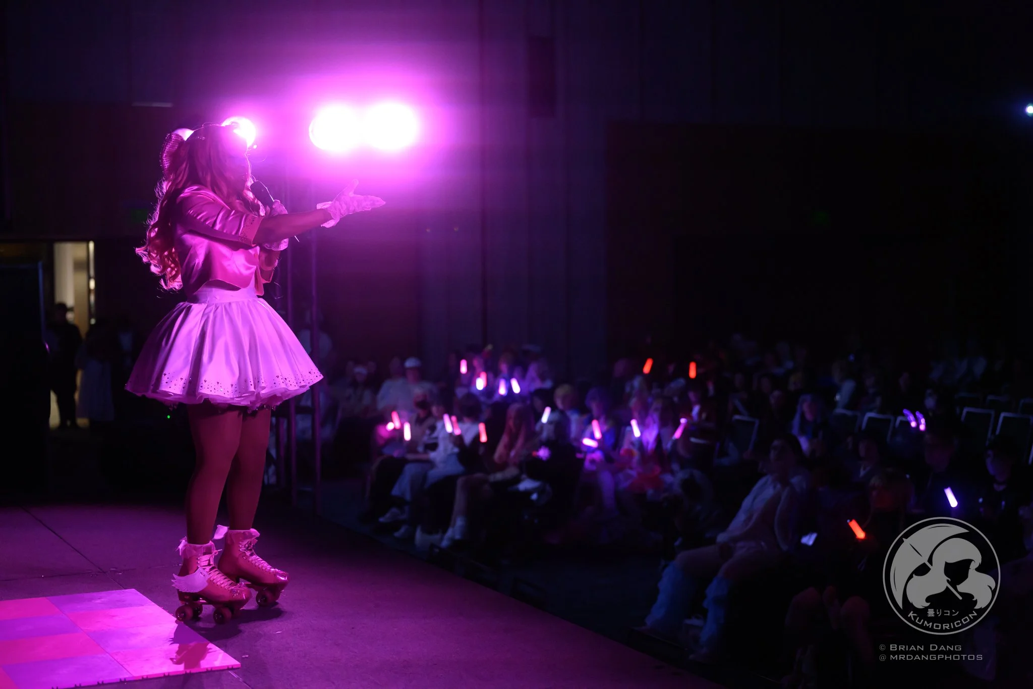 A performer dressed in pink with roller skates, on stage at a dark venue, speaking to an audience with glow sticks.