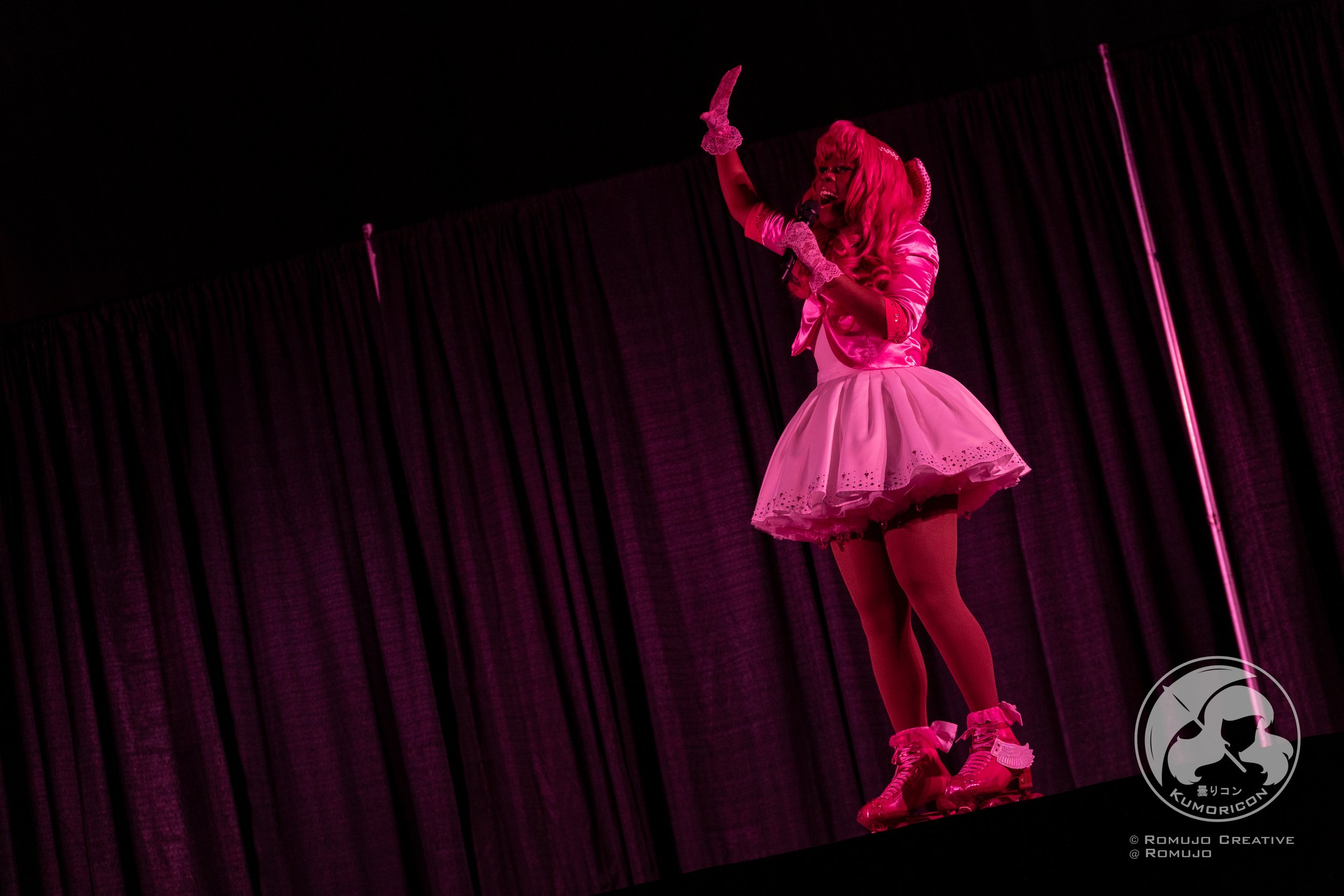 Performer dressed in pink costume on stage with dark curtain backdrop, holding microphone and gesturing passionately.
