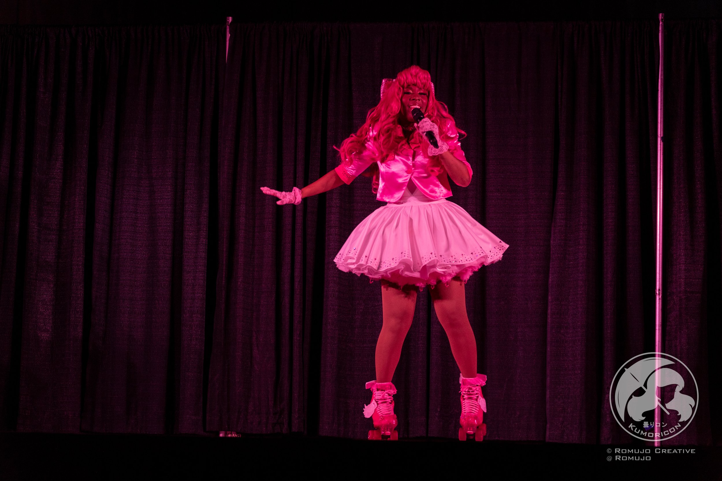 Performer on stage wearing a pink outfit with roller skates, singing into a microphone with a black curtain backdrop.