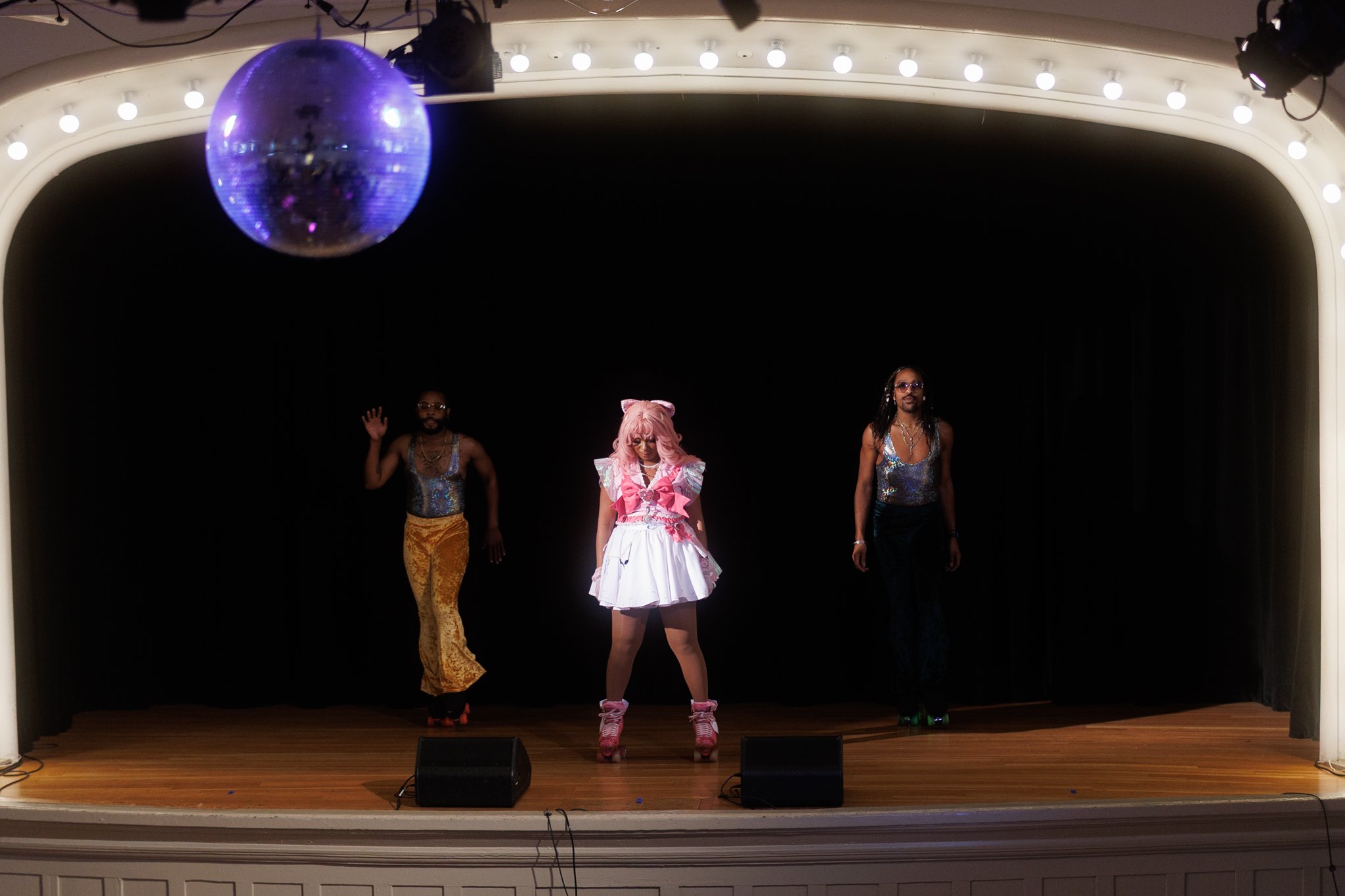 A performer dressed in pink and white with a pink bow and roller skates stands center on stage, flanked by two dancers in colorful outfits, under a disco ball and stage lights with a black curtain background.