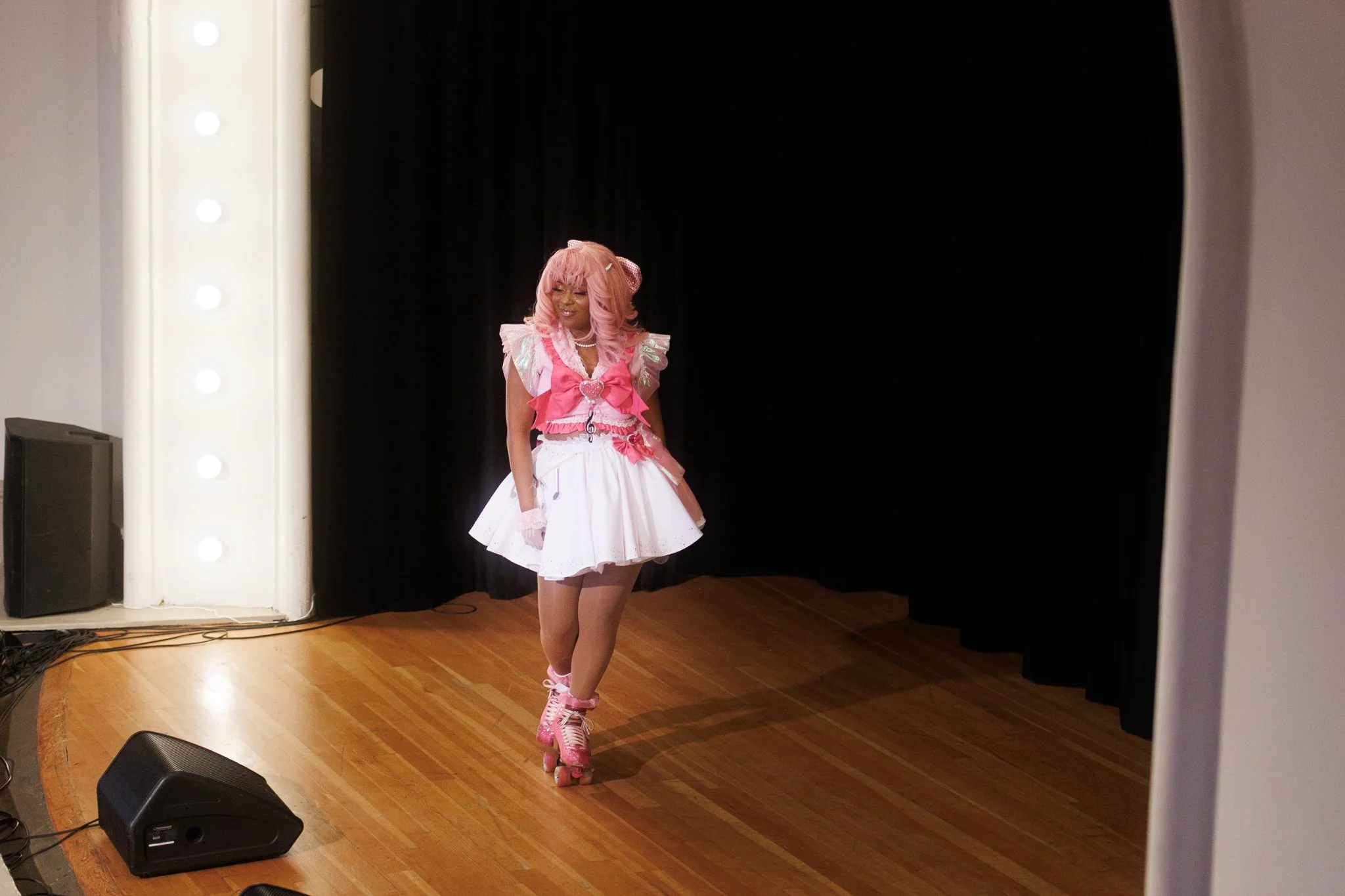 A woman dressed in a pink and white costume on stage with black curtains, with a wooden floor and stage lights in the background.