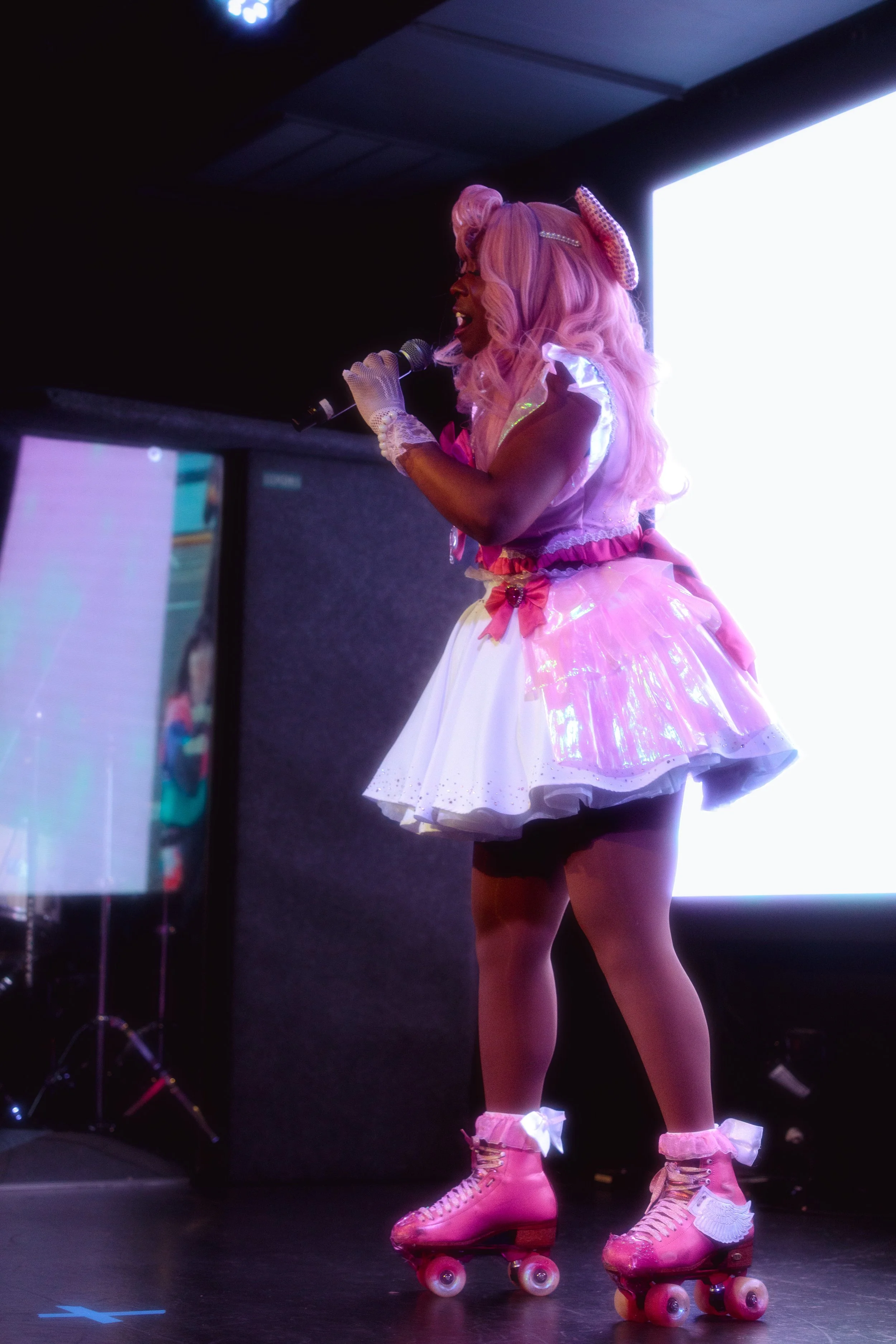 A woman dressed in a pink and white lolita-style costume with pink roller skates, singing into a microphone on a stage with bright screens in the background.