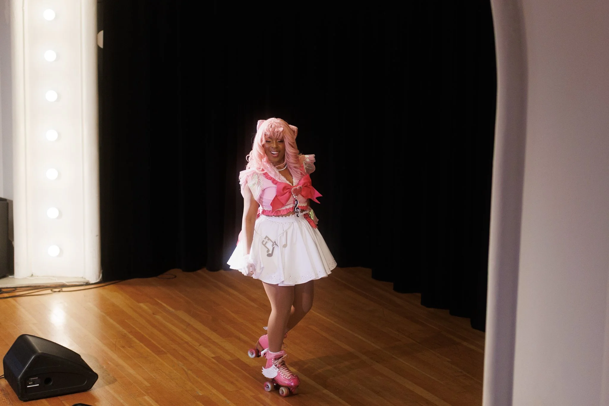 A person dressed in a pink and white outfit, including a skirt and a bow, roller skating on a wooden floor stage with black curtains and a lit mirror with bulbs nearby.