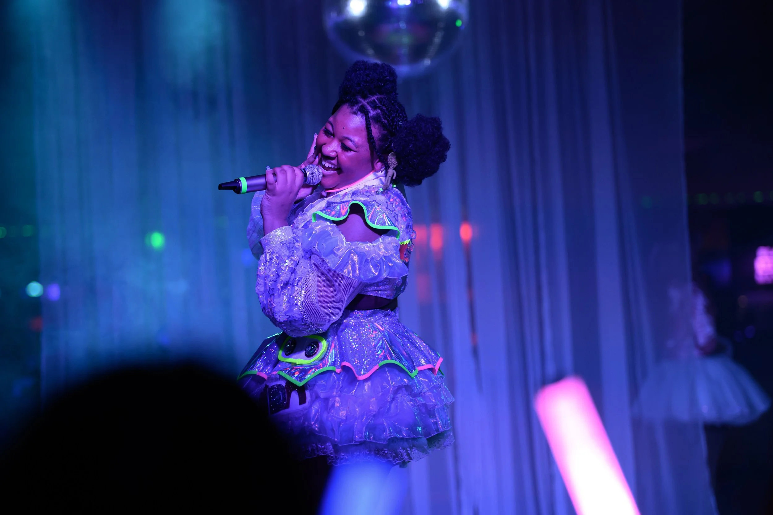 A woman singing or speaking into a microphone on a stage illuminated with vibrant, colorful lighting, wearing a shiny, purple dress with neon accents. She appears joyful, with her hand on her face, and her hair styled in two large puffs.
