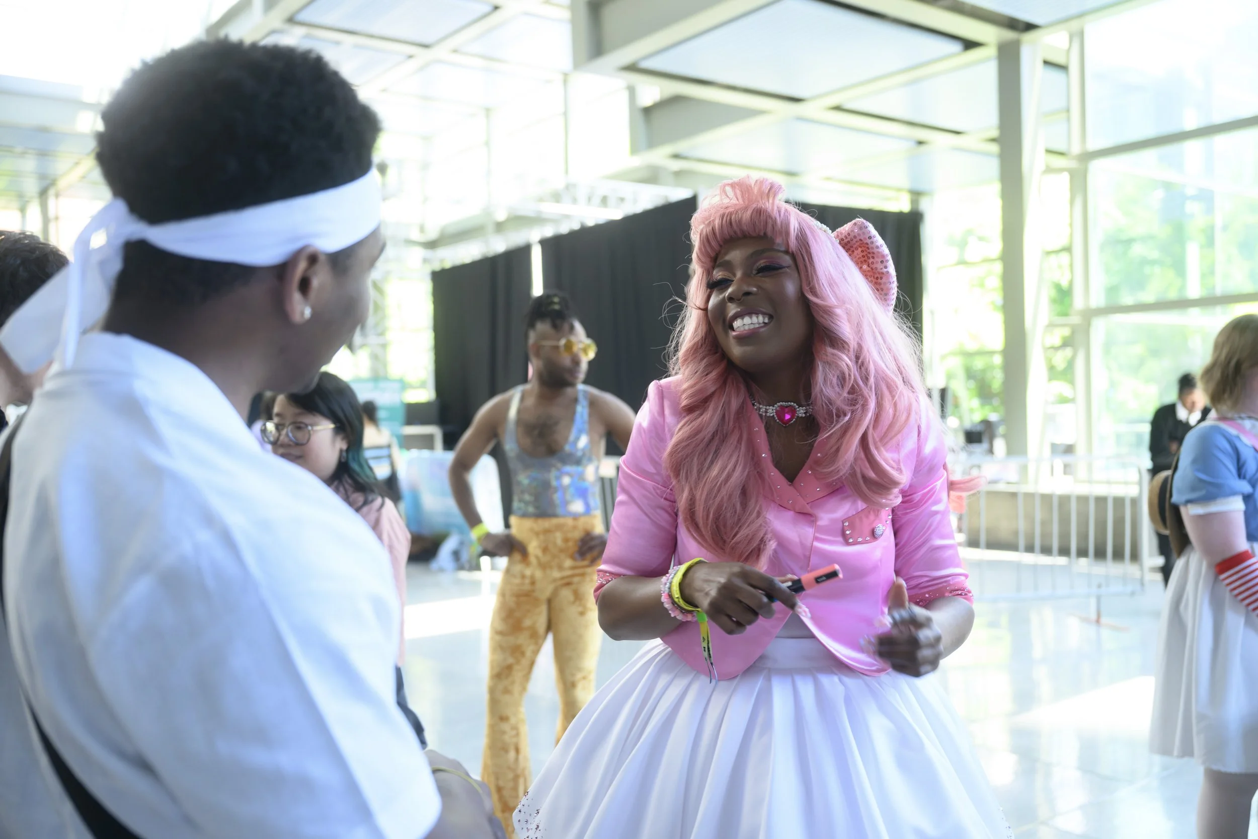 A woman with pink hair and a pink jacket smiling and talking to a man in a white shirt at an indoor event.