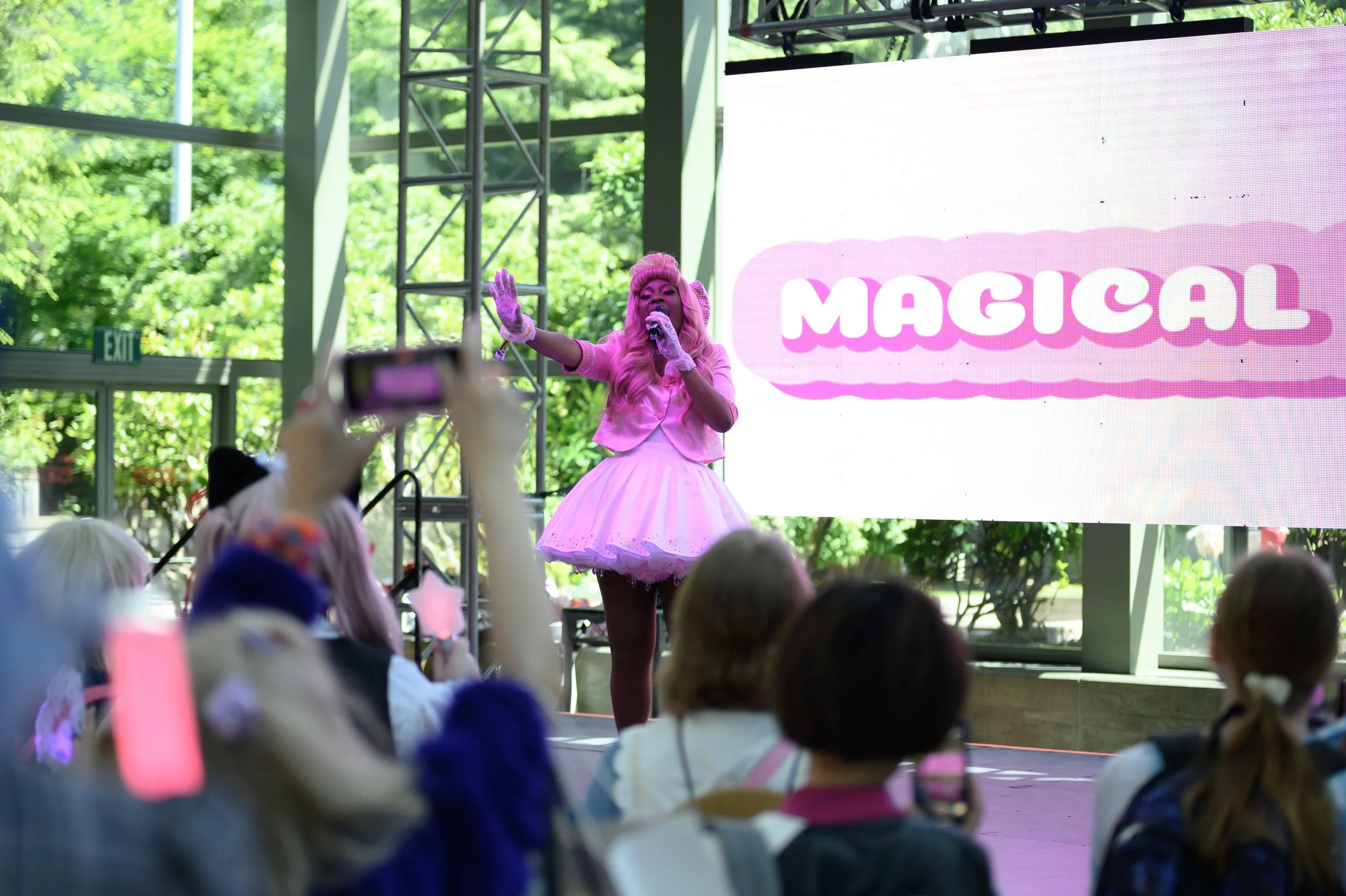 Performer dressed as a pink princess singing on stage at an event, with a large screen displaying the word 'MAGICAL' in the background and an audience taking pictures.