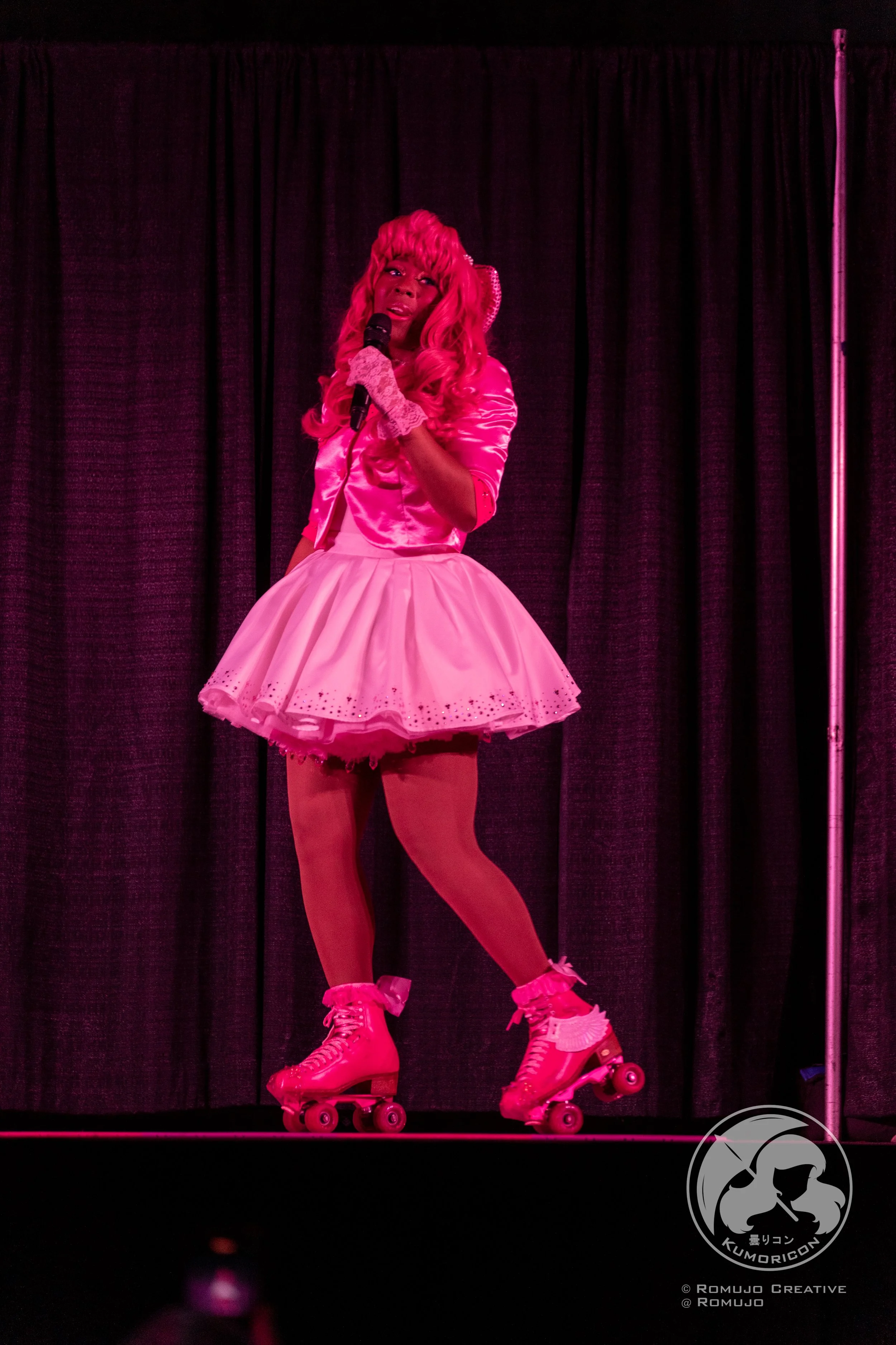 Person dressed in pink, wearing roller skates, standing on stage with black curtains, holding a microphone.