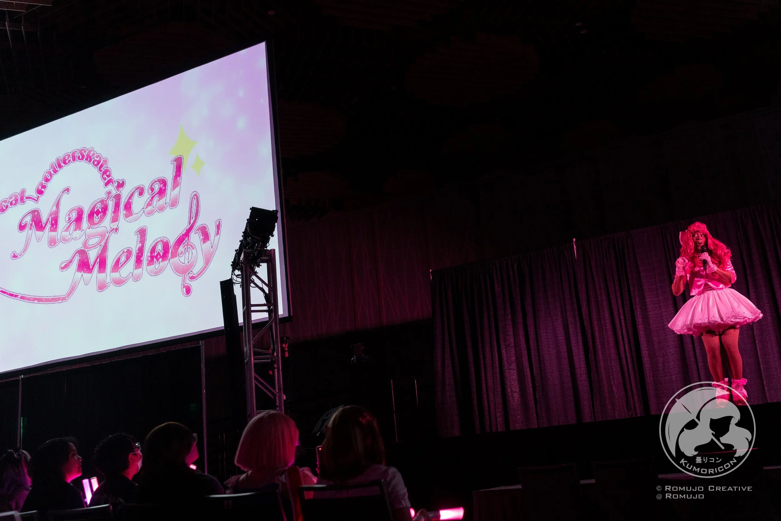 A stage performance at a convention with a person dressed in pink, wearing a tutu and wig, speaking into a microphone. A large screen displays the logo 'Magical Melody,' and an audience watches from the darkened room with pink lighting.