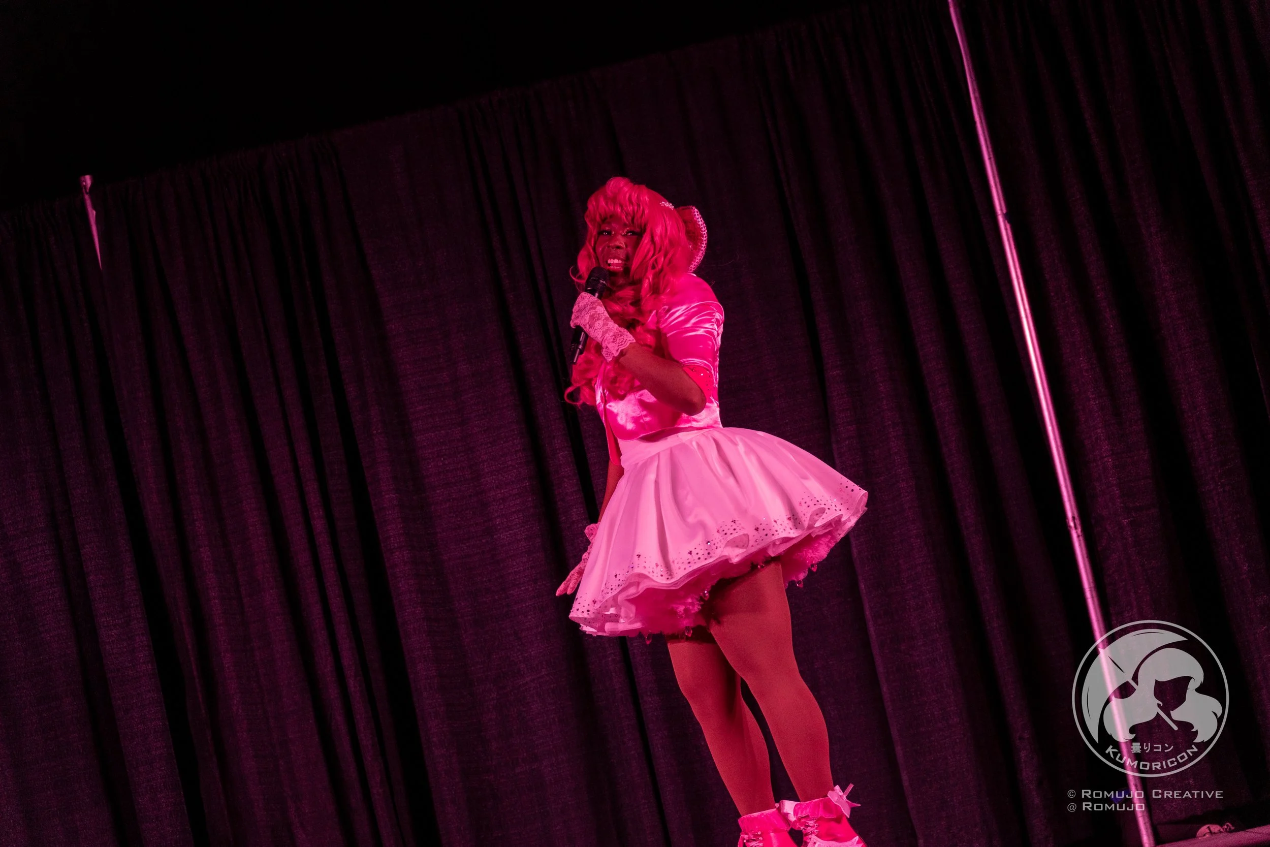 Performer dressed in pink outfit, wig, and pink roller skates on stage with black curtain background.