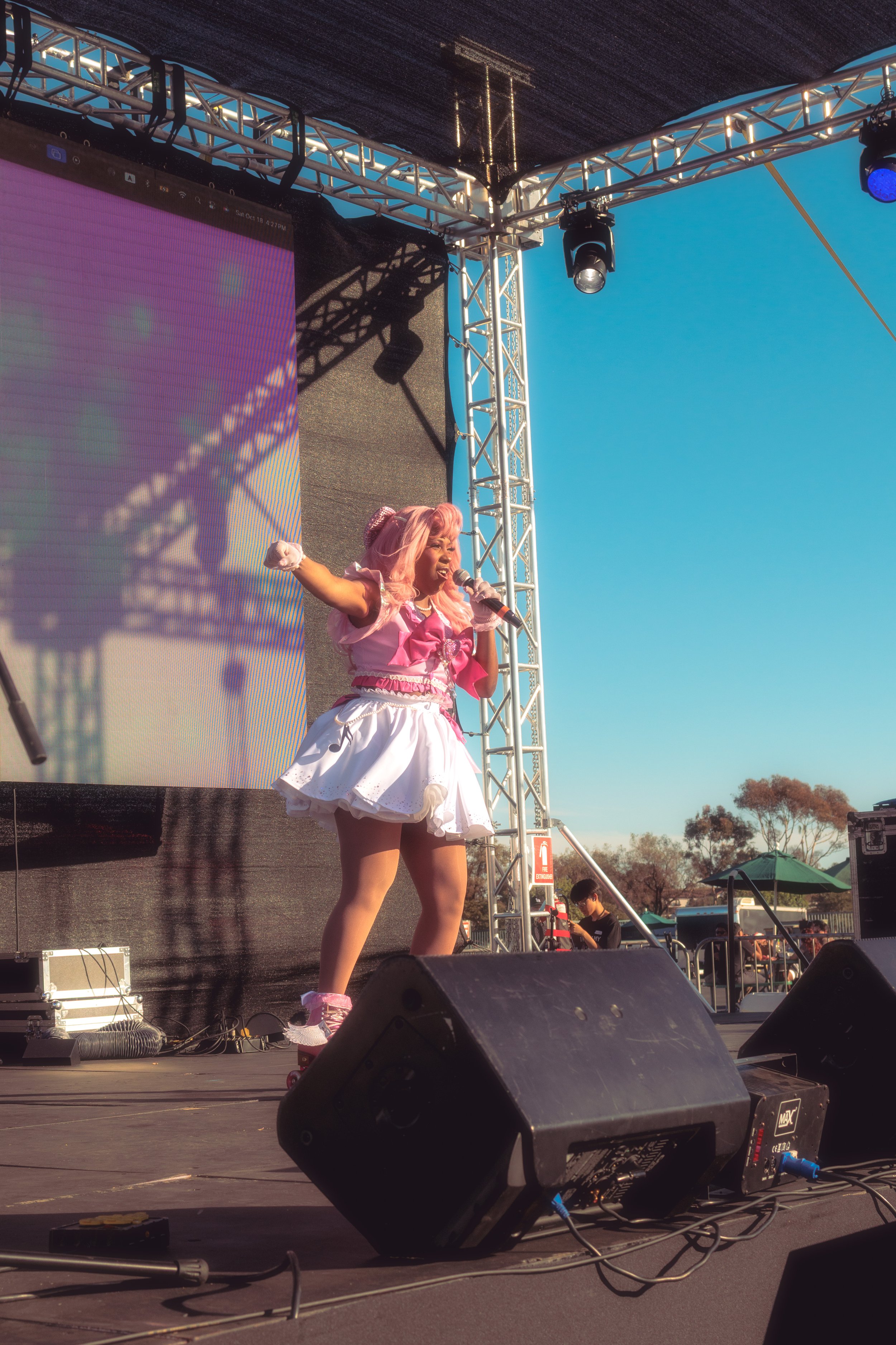 Performer dressed in pink and white costume singing into a microphone on an outdoor stage with a large screen and metal truss overhead, during daytime.