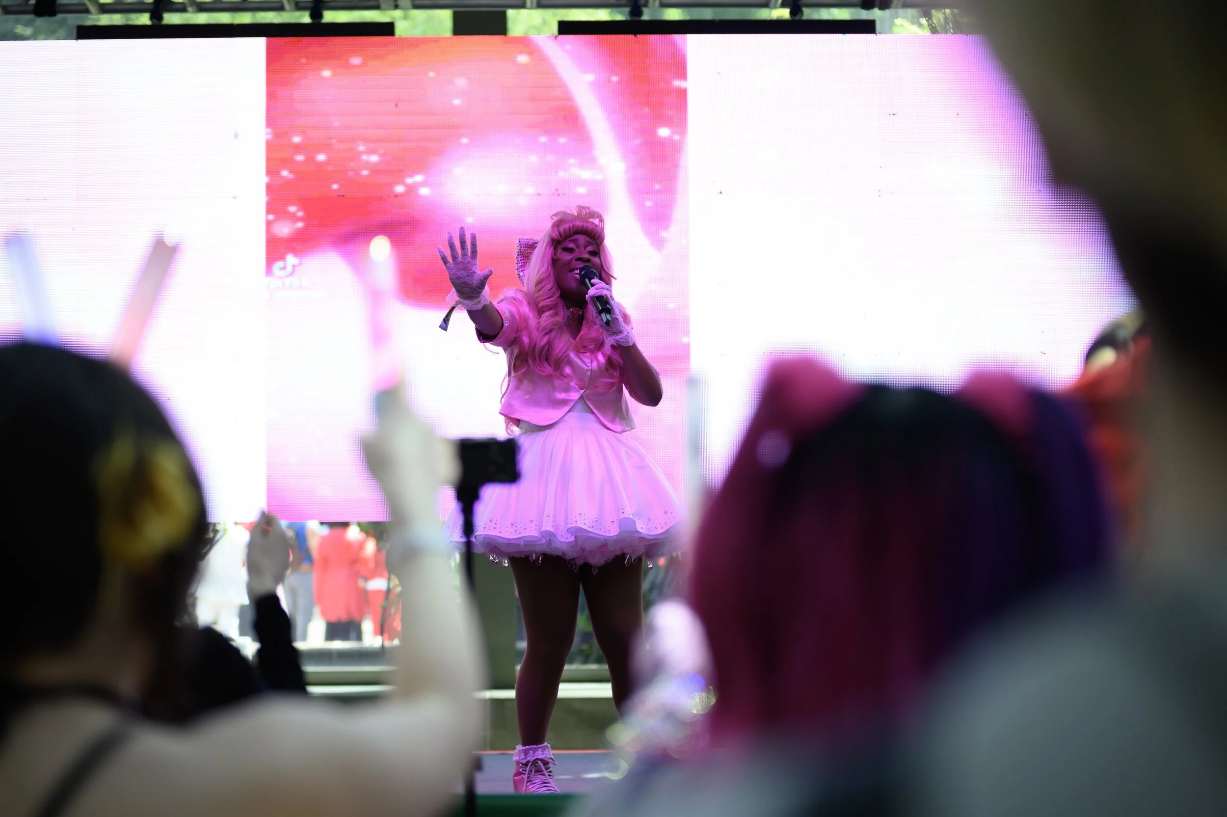 Performer dressed in pink, holding a microphone on stage with a large pink and white digital screen background.