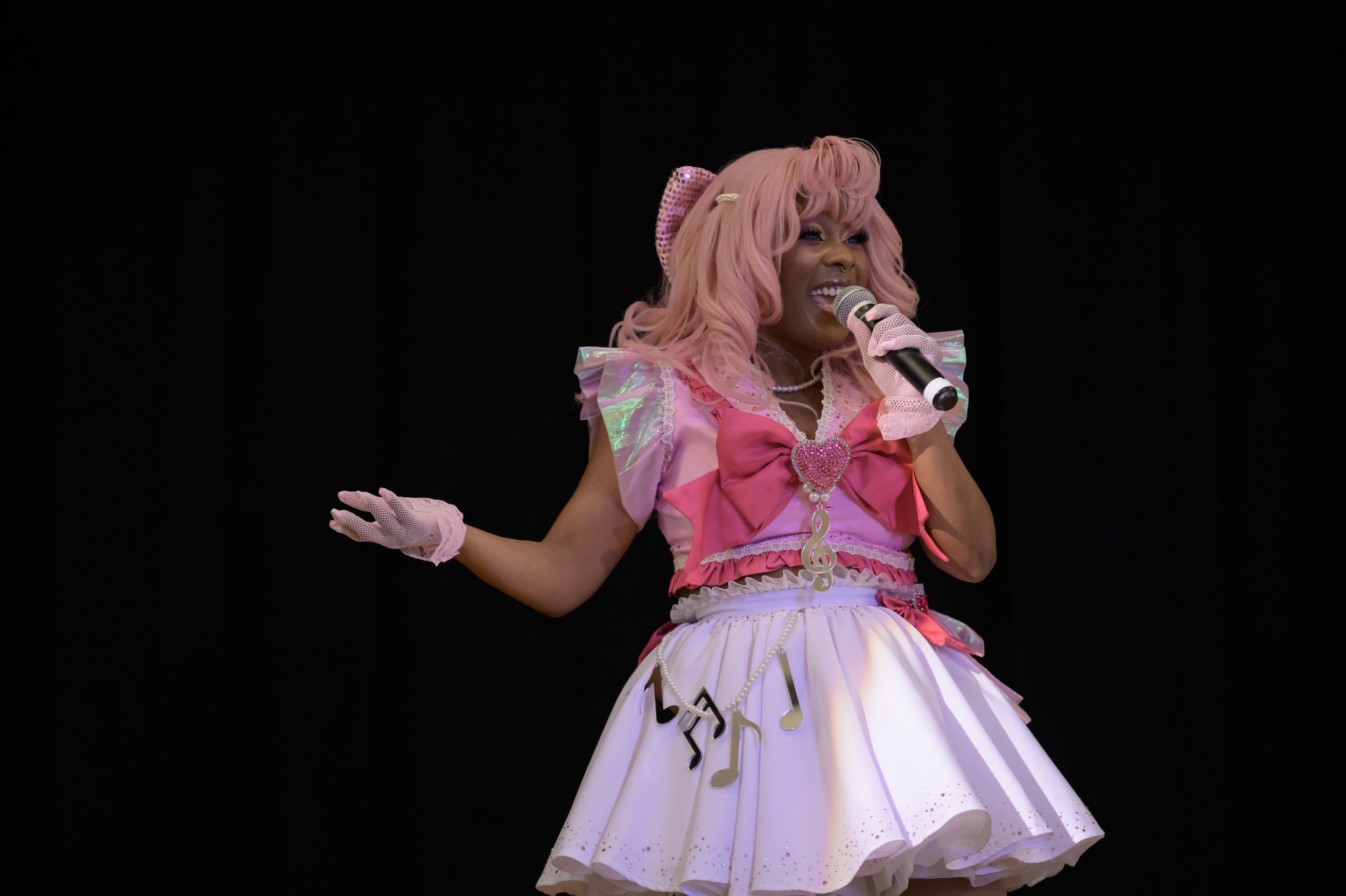 A woman dressed in a pink and white costume with musical notes hanging from her skirt, pink hair, and pink gloves, holding a microphone and singing or speaking on stage.